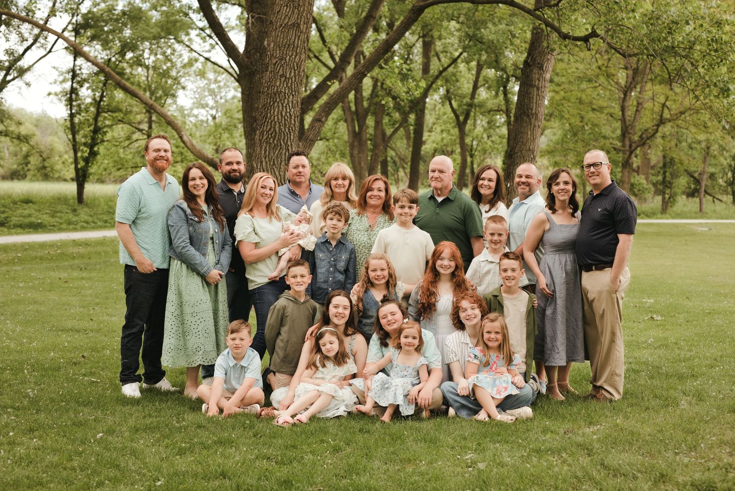 Large extended family group portrait outdoors in a lush green park, posing together under a large tree.