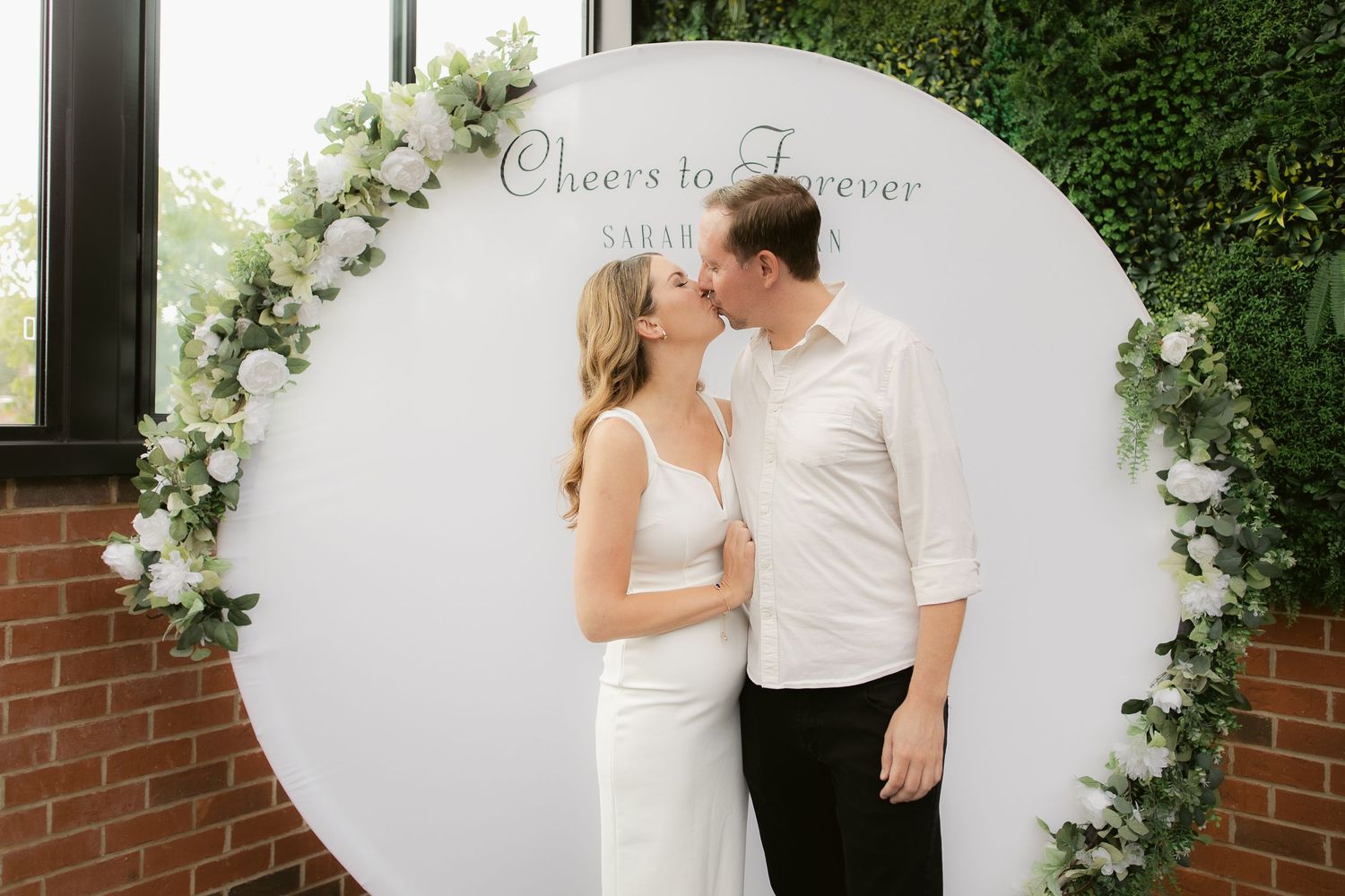 Couple kissing in front of a circular floral arch with a Cheers to Forever sign at an elegant engagement party.