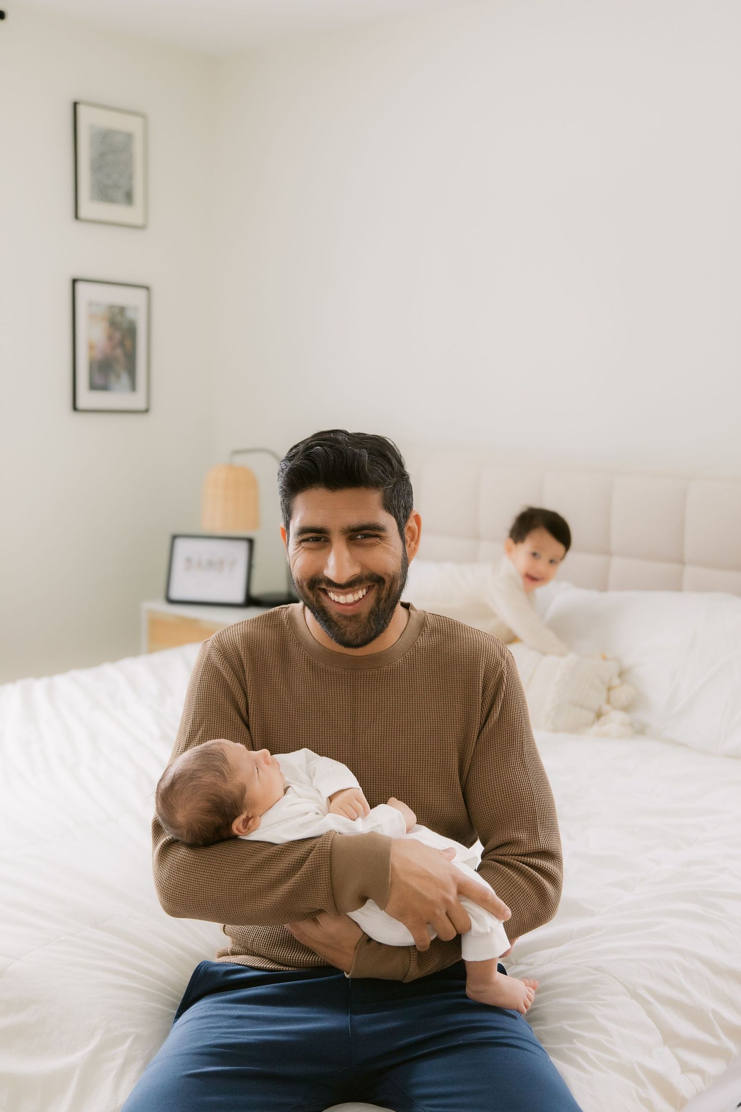 Parents share a tender moment with their newborn baby on a white bed with framed artwork on the wall.