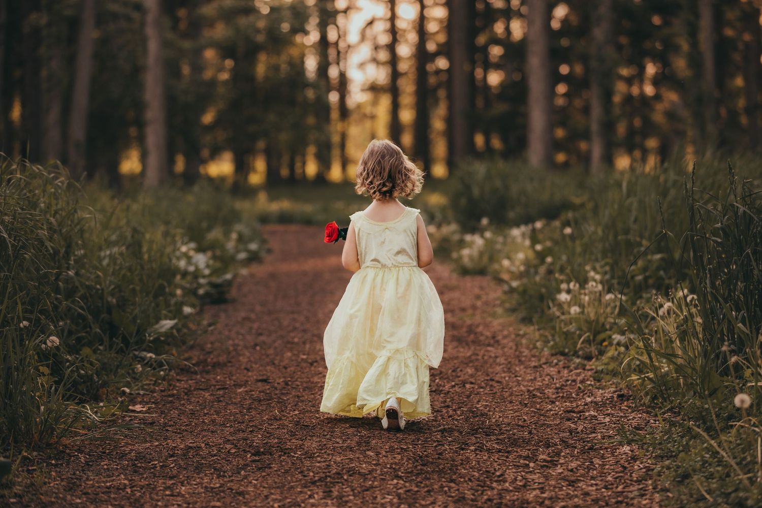 Young girl in yellow dress walks down forest path surrounded by wildflowers at sunset.