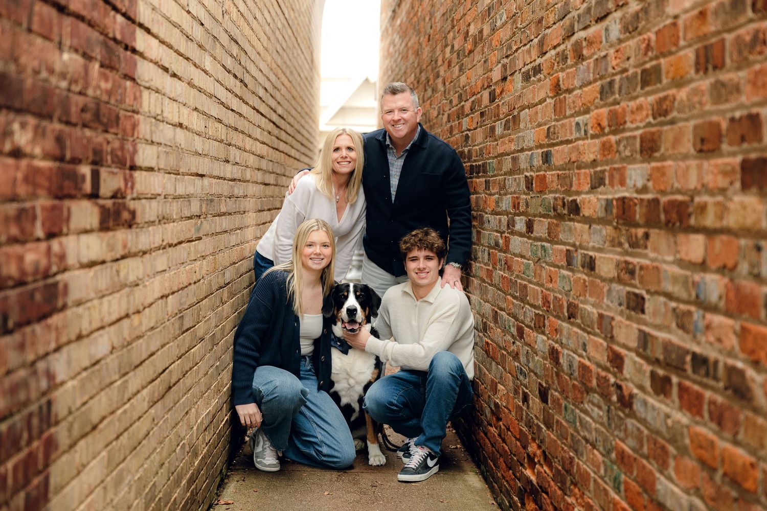 Family and pet dog pose together in narrow brick alleyway setting.