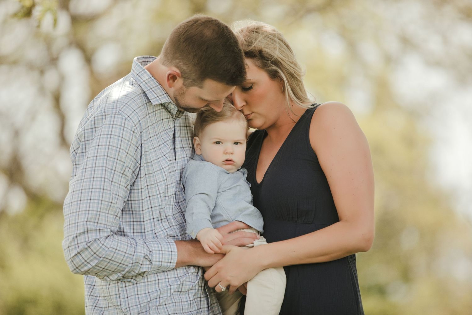 A couple tenderly holds their baby boy outdoors during a family portrait session in a park setting.