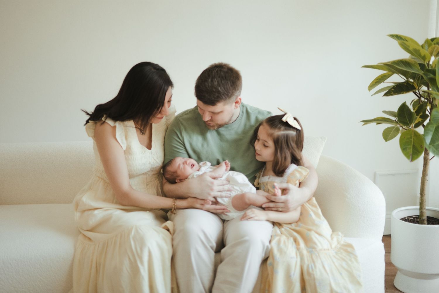 Family of four sitting on a white sofa, parents lovingly gazing at newborn baby with young daughter nearby.