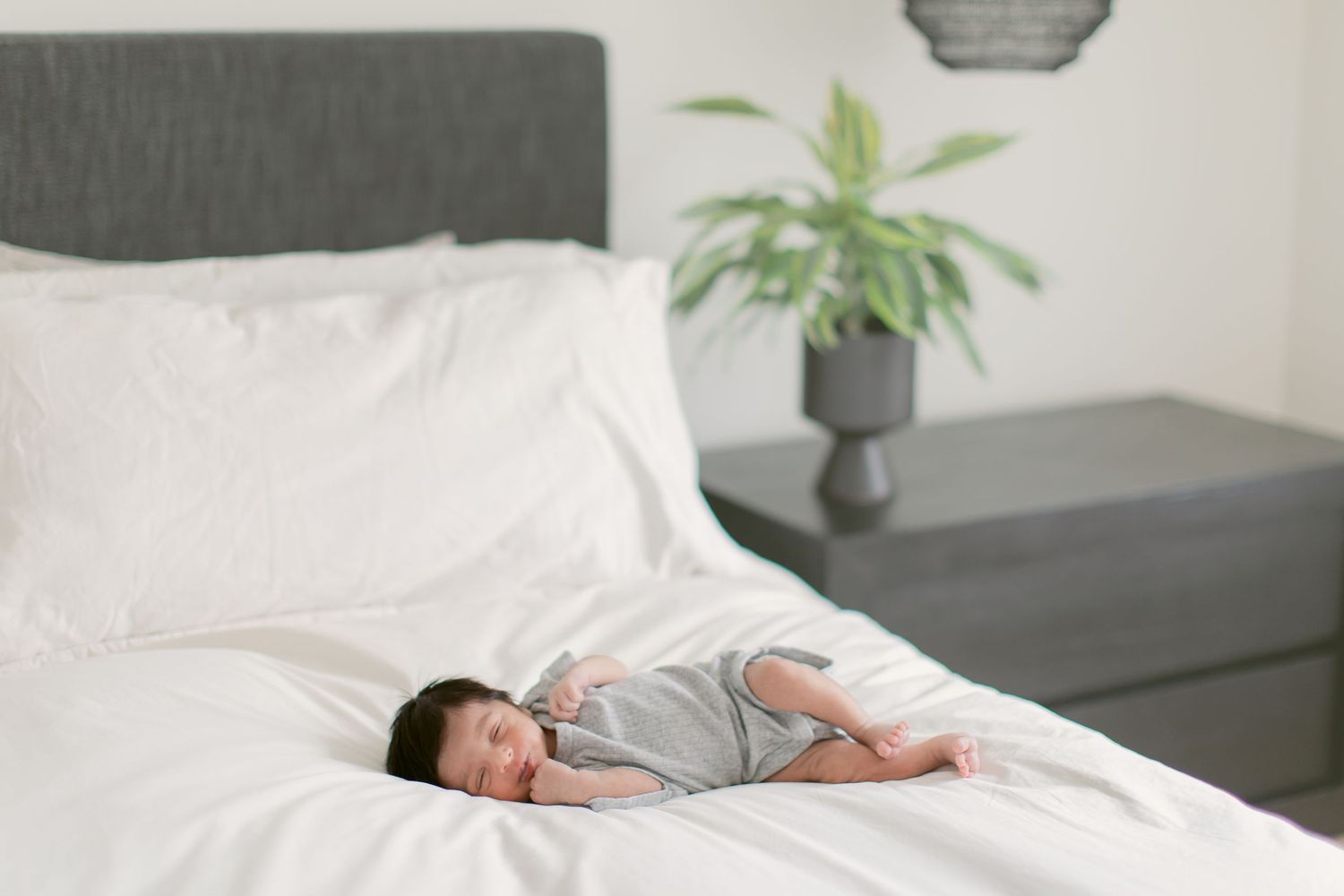 Peaceful newborn baby in gray onesie sleeping on white bedding near a modern gray headboard and green plant.