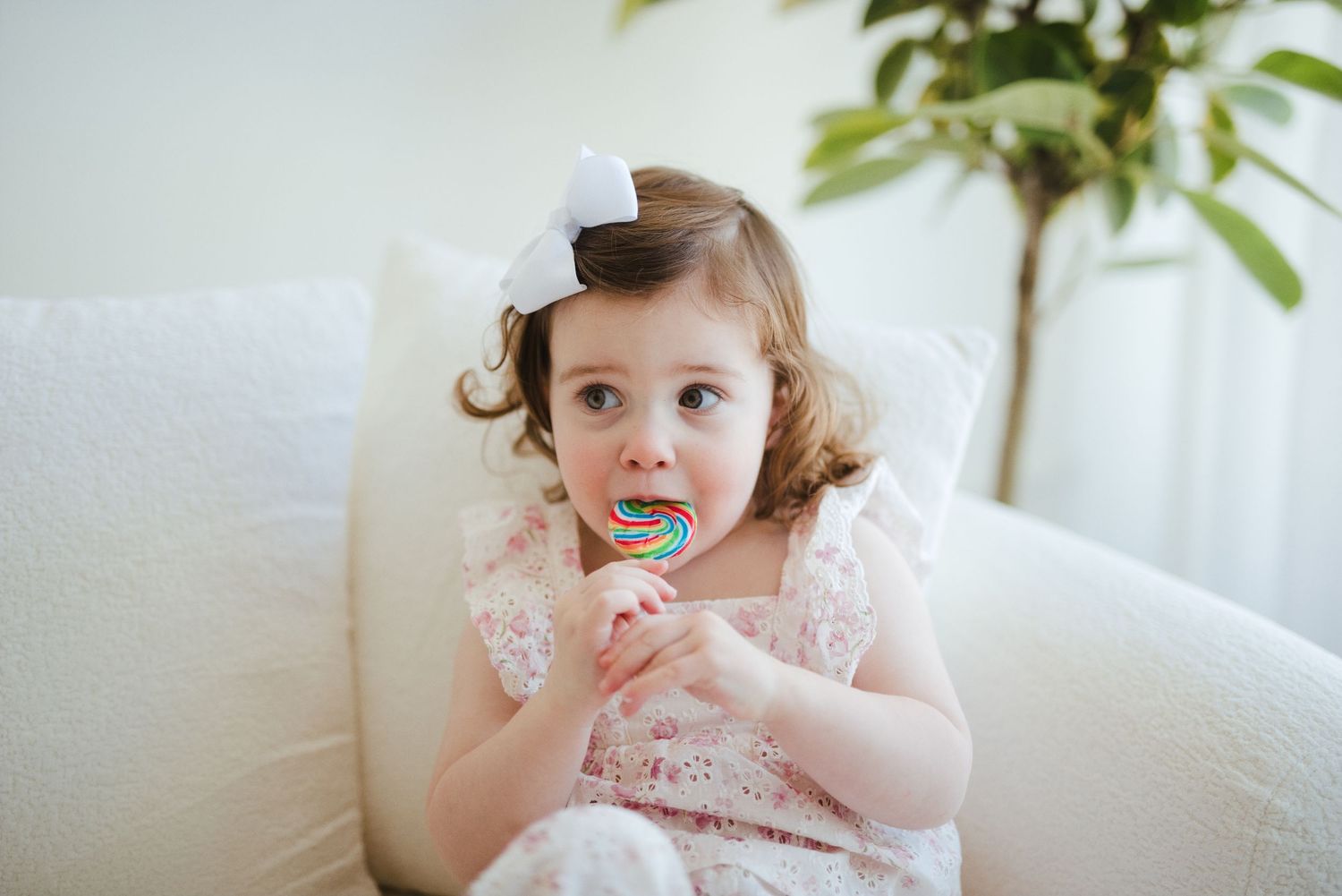 Adorable toddler girl with white bow eating colorful lollipop on white chair in bright room.