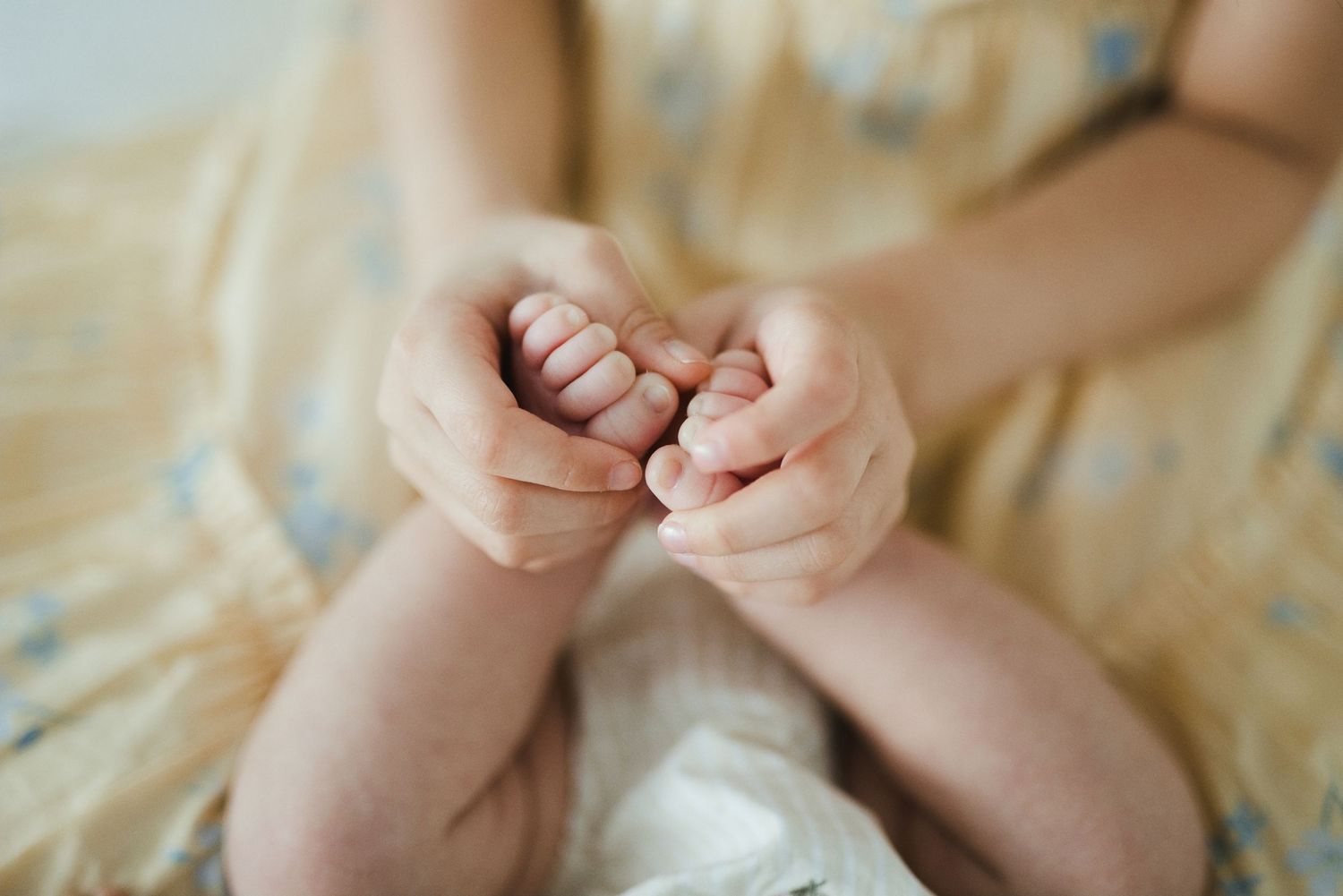 Close-up of a baby's tiny hands gently clasped together, resting on a soft yellow floral fabric.