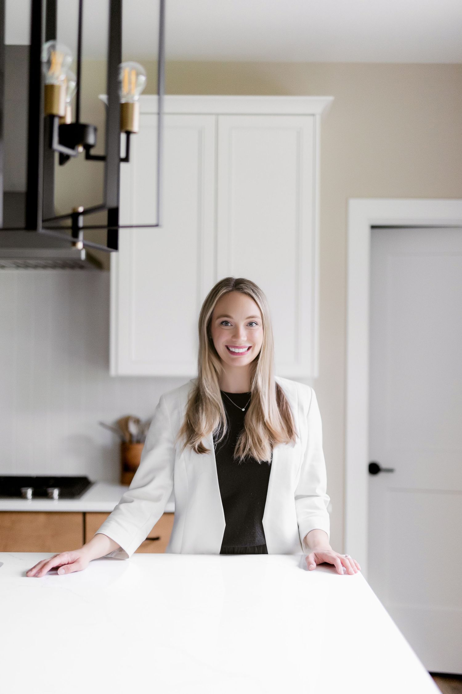 Professional in white blazer stands at kitchen counter under modern pendant lighting.