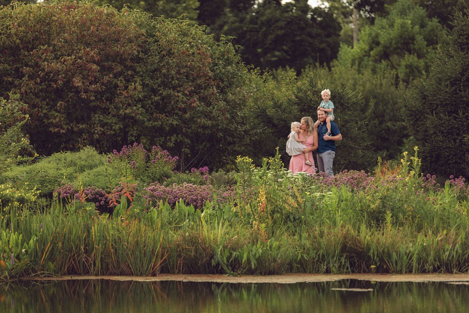 English country garden pond with wooden bridge and lush summer plantings surrounded by trees and foliage.