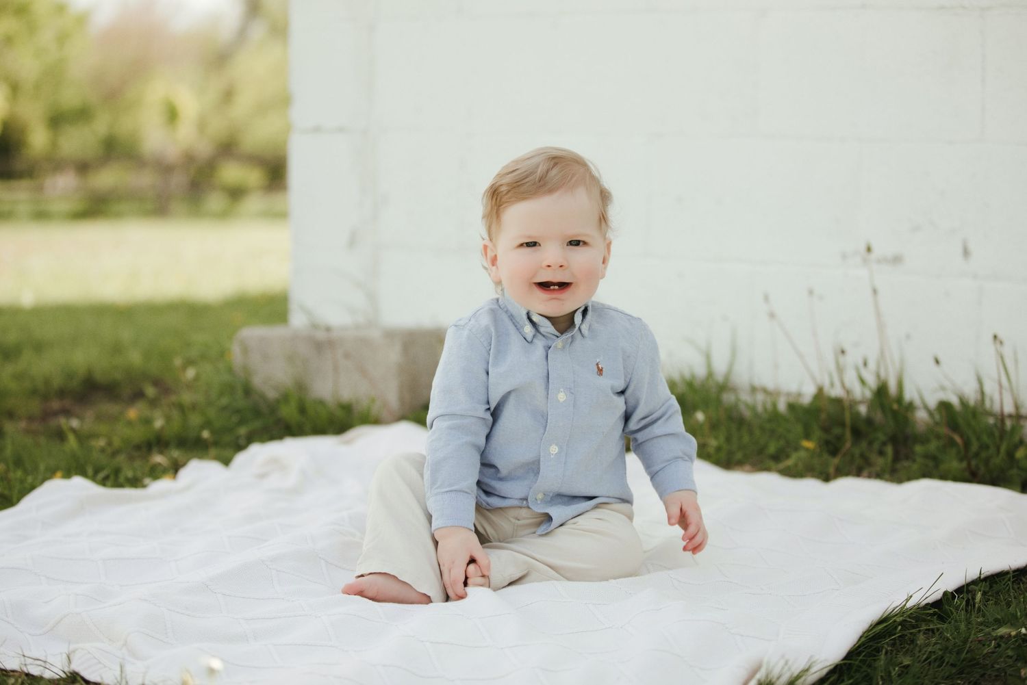 A happy baby boy in a blue shirt and khaki pants sits on a white blanket outdoors on green grass.