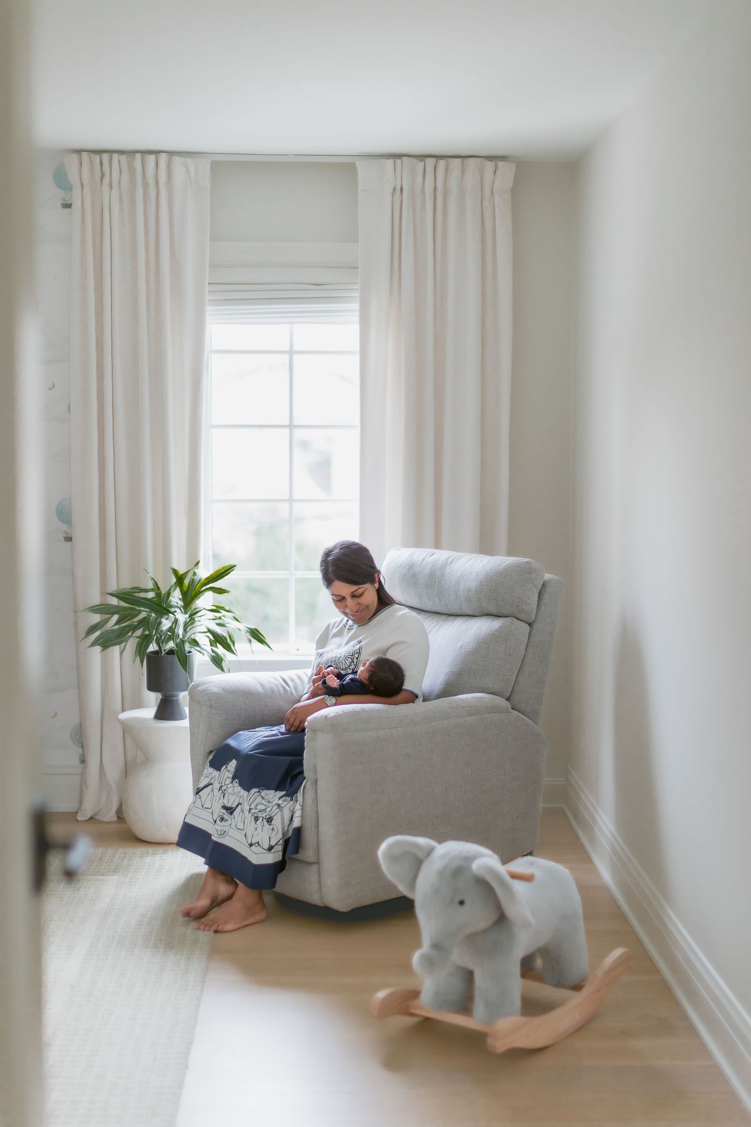 A mother nursing her newborn in a gray glider chair in a bright nursery with an elephant rocker nearby.
