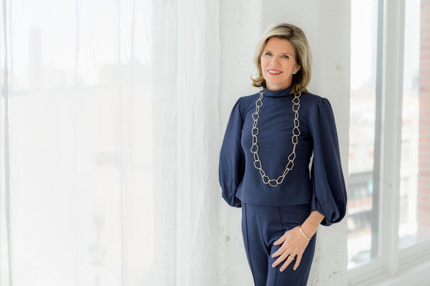 Professional headshot of someone in a navy blue outfit with statement necklace against white background.