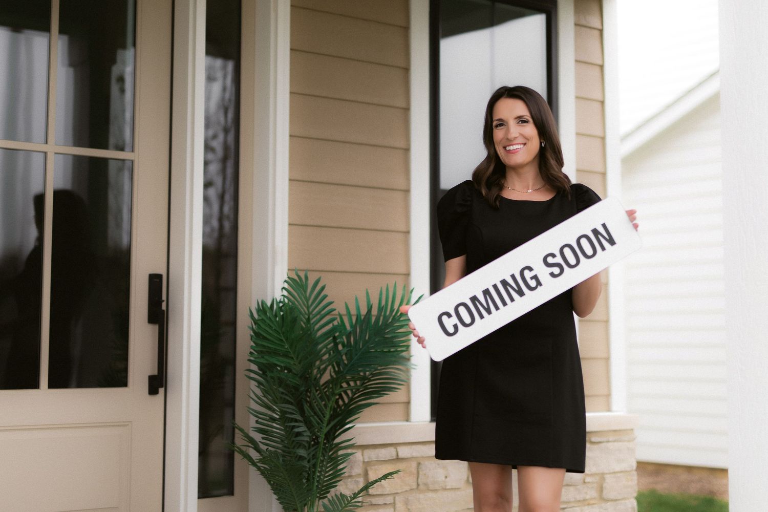 Person in black dress holding Coming Soon sign outside tan house with potted palm.