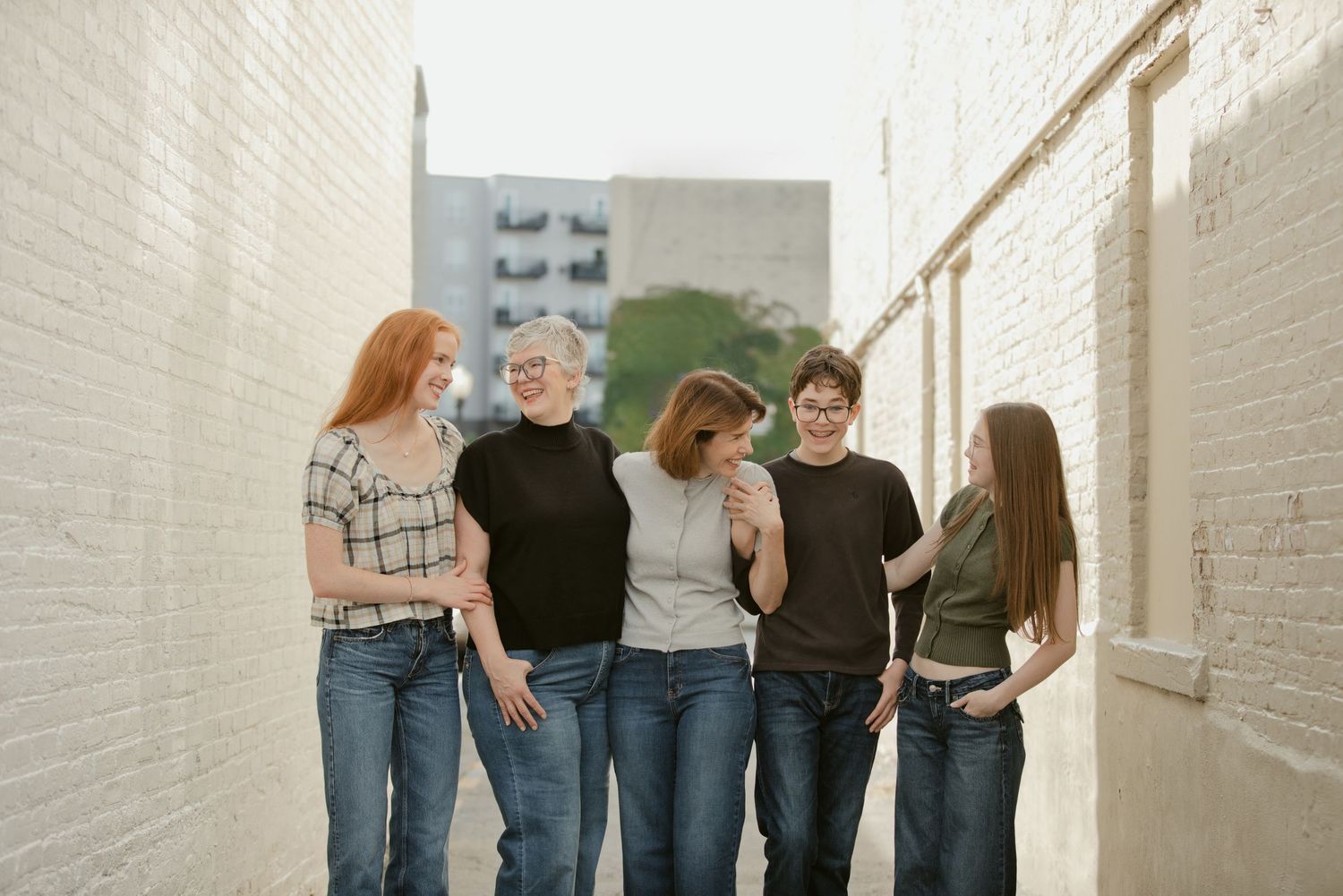 Five women laughing and walking together in an urban alleyway between white brick walls.