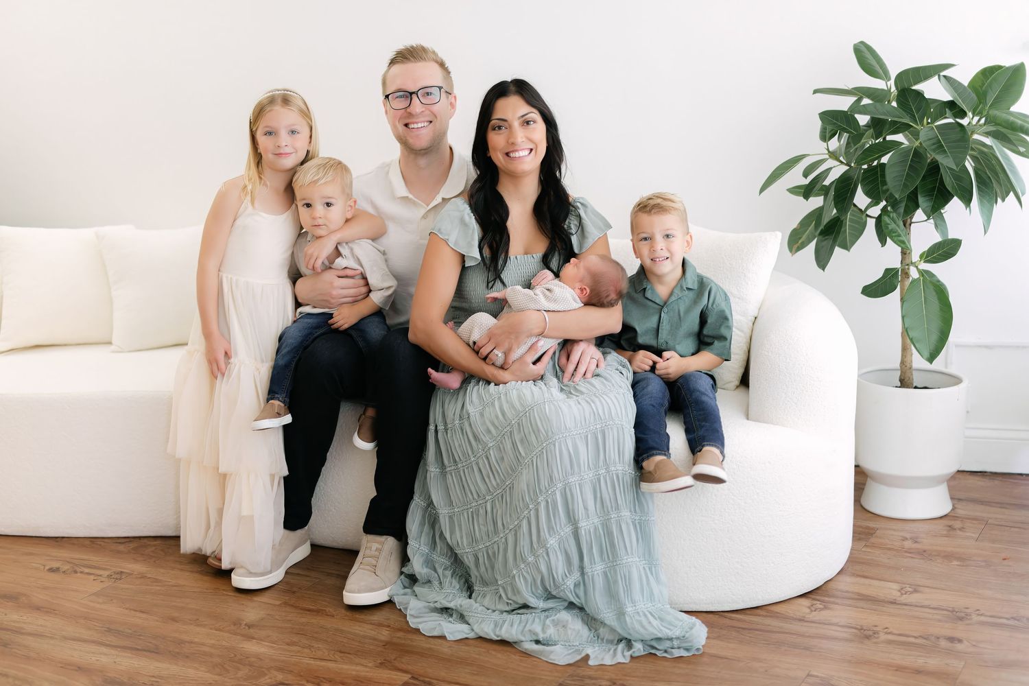 A happy family of six poses together in a bright white studio, including a newborn baby, on a modern white sofa.
