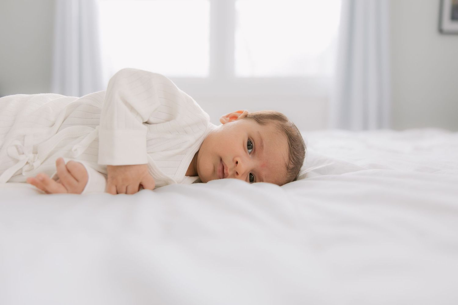 A person wearing white lies comfortably on crisp white bedding in a bright, sunlit bedroom.