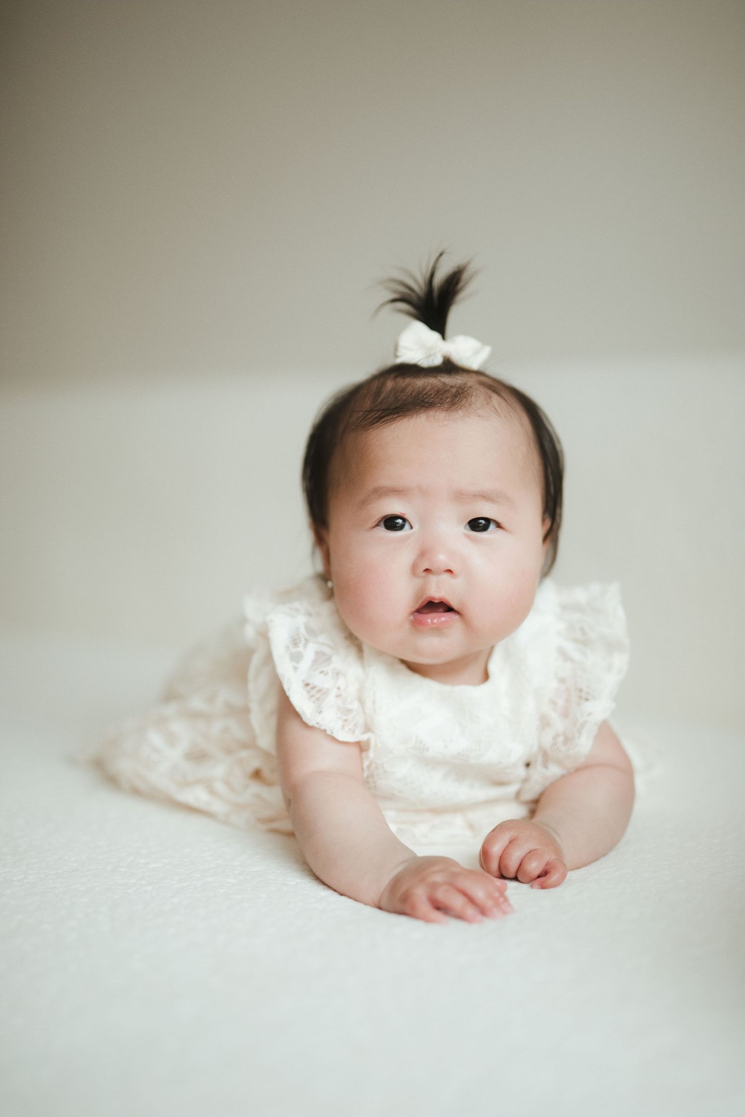 Adorable baby girl in white lace romper with tiny hair bow lying on white surface, looking curiously at camera.