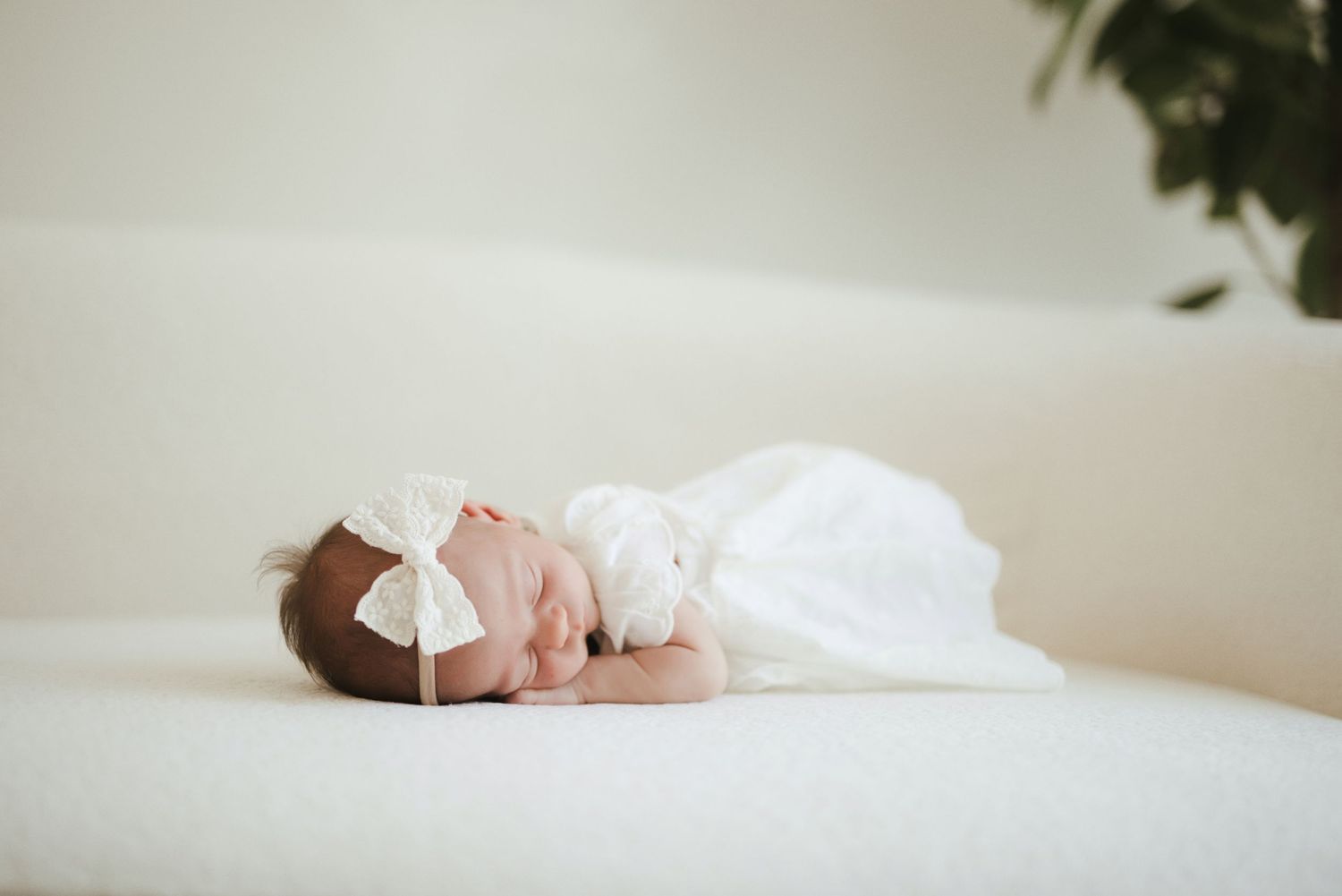 Newborn baby sleeping peacefully in white swaddle with lace bow headband on soft white surface.