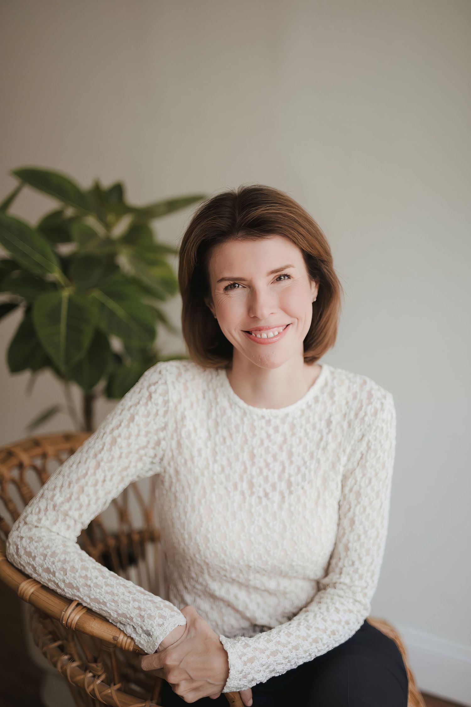 Smiling woman in white knit sweater sitting in wicker chair with green plant in bright modern room.