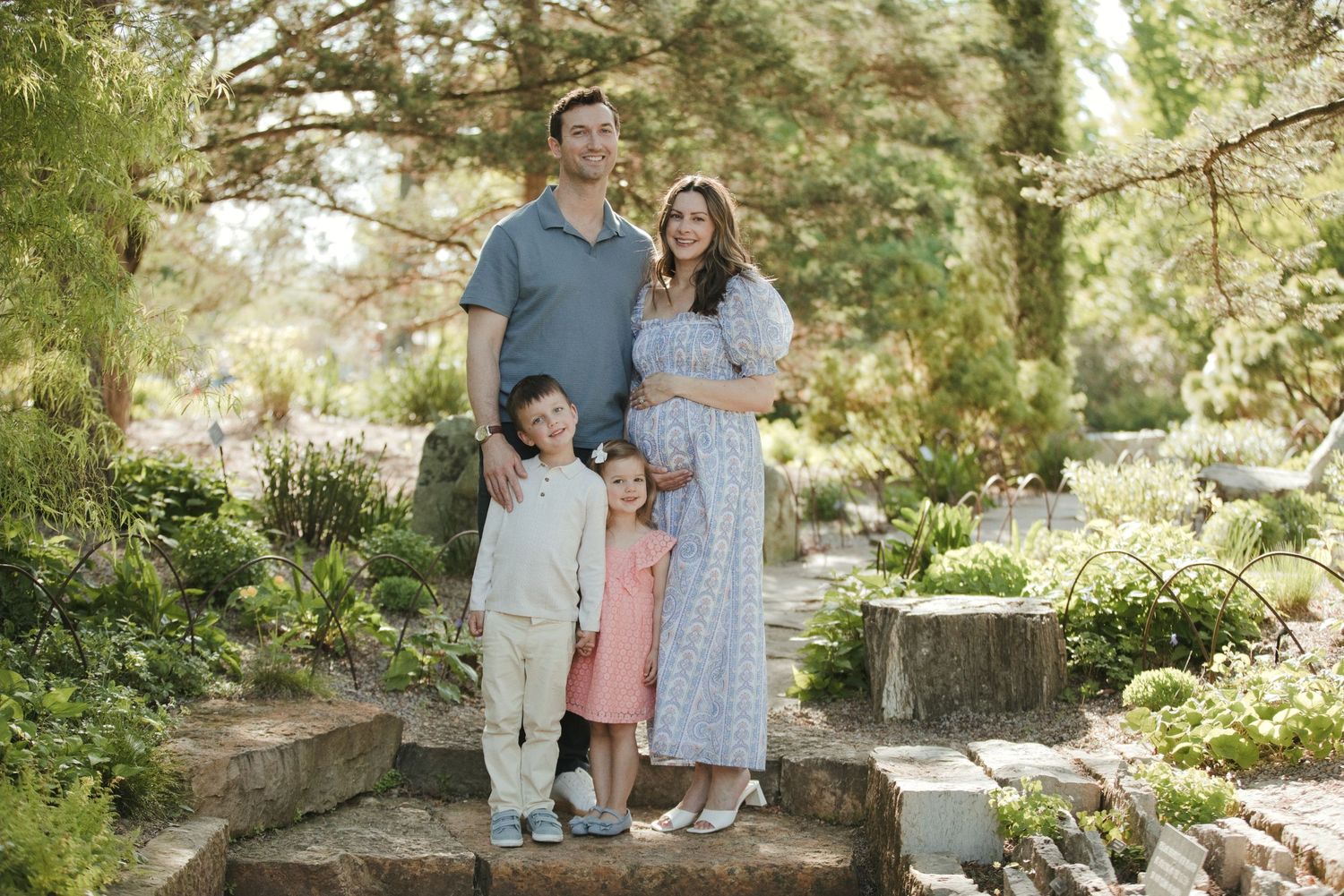 Family of four poses on stone steps in a lush garden, with parents and two young children smiling outdoors.