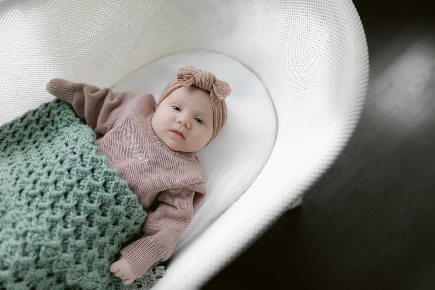 Newborn resting in white bassinet with mint green knit blanket.