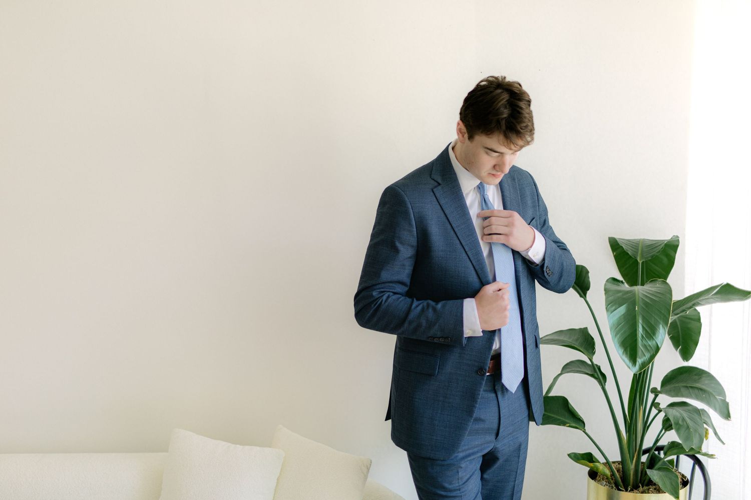 A young man in a navy blue suit adjusts his tie while standing near a white sofa and green plant.