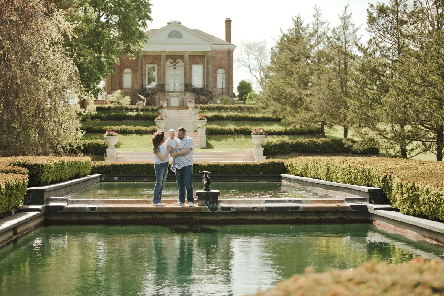 Couple standing by a reflecting pool in a formal garden with terraced hedges and a brick mansion backdrop.