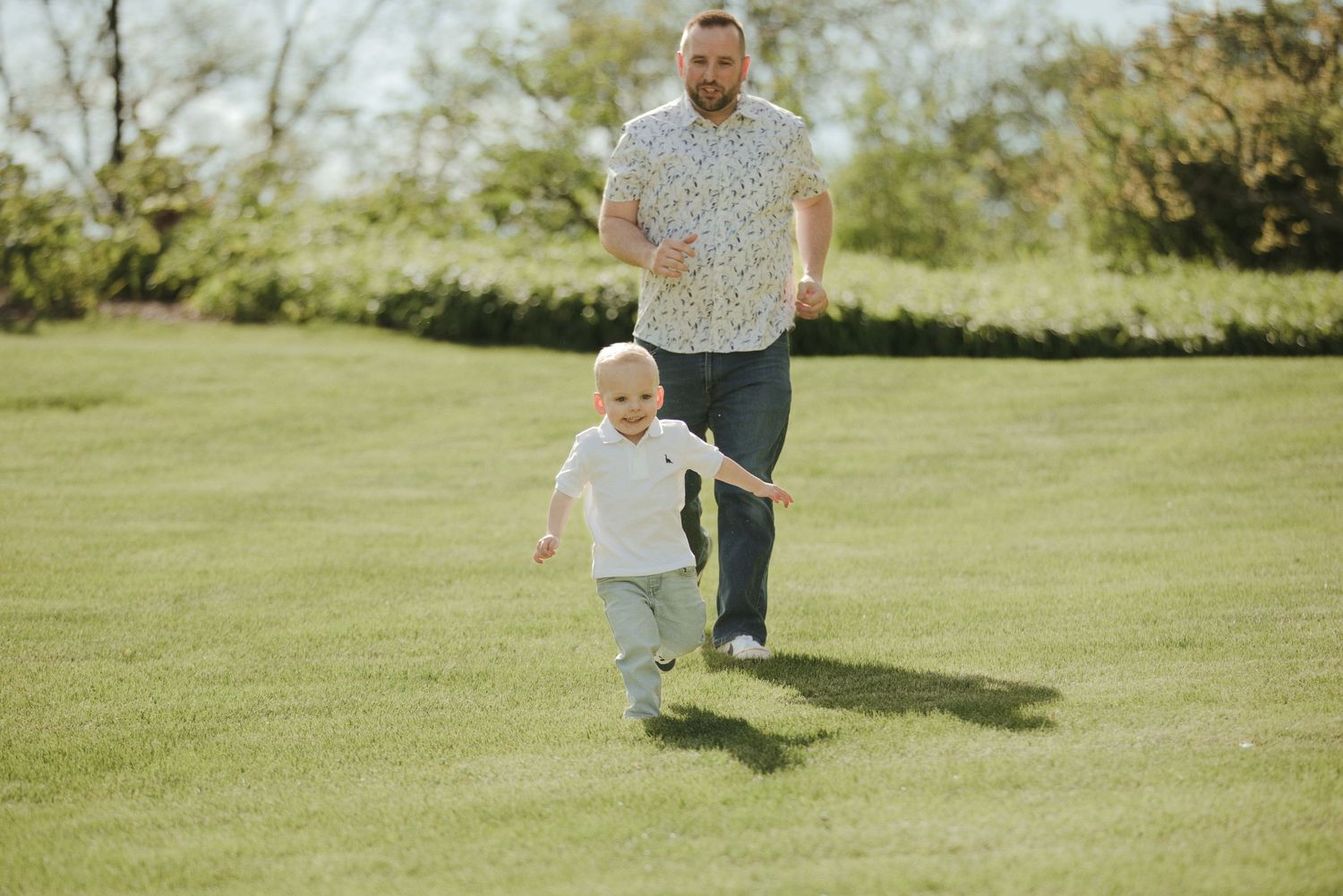 Father chasing happy toddler across a green lawn in a sunny outdoor park setting.