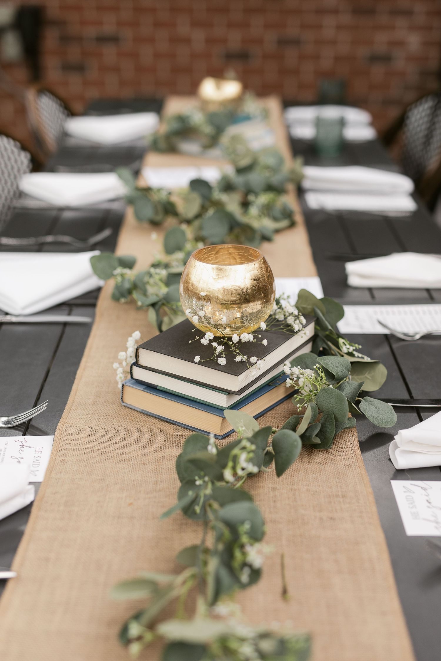 Elegant wedding table centerpiece with stacked books, gold candle holder, eucalyptus garland, and white napkins on dark wood.