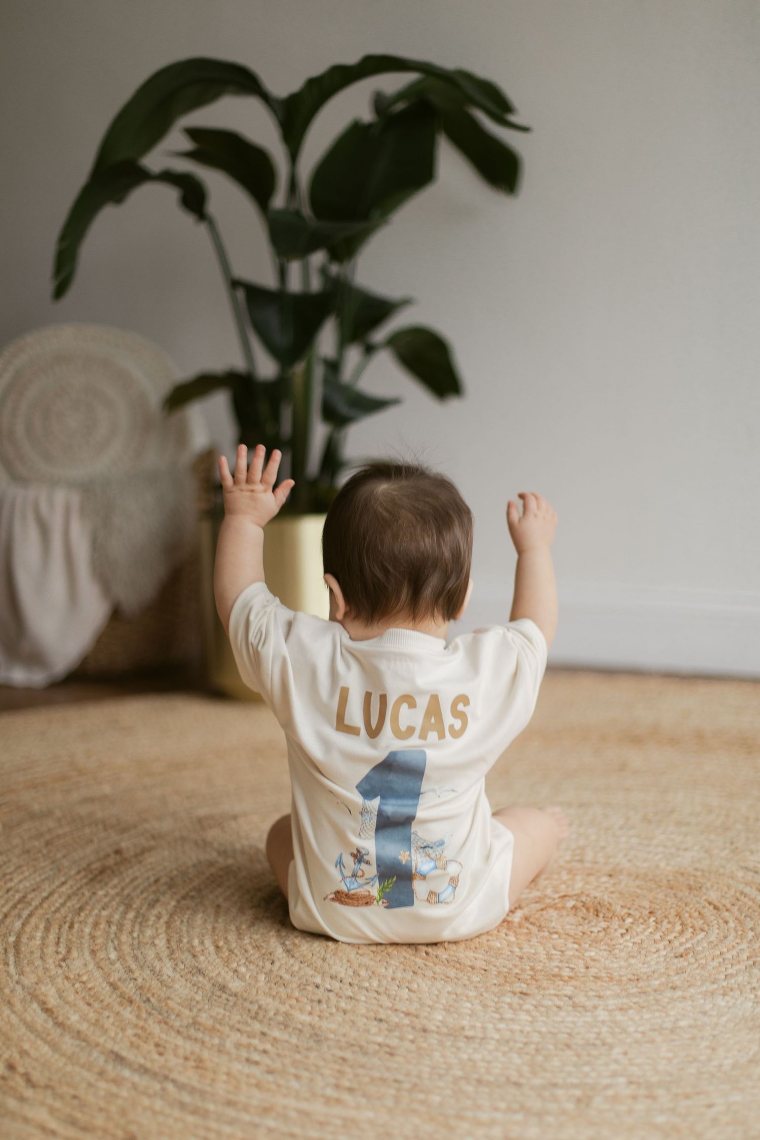 Baby wearing white shirt with name and number 1 sits on round jute rug with arms raised near indoor plant.