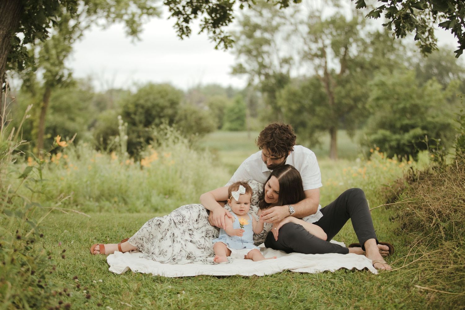 Young family with baby relaxing on white blanket in lush green park during outdoor photo session.