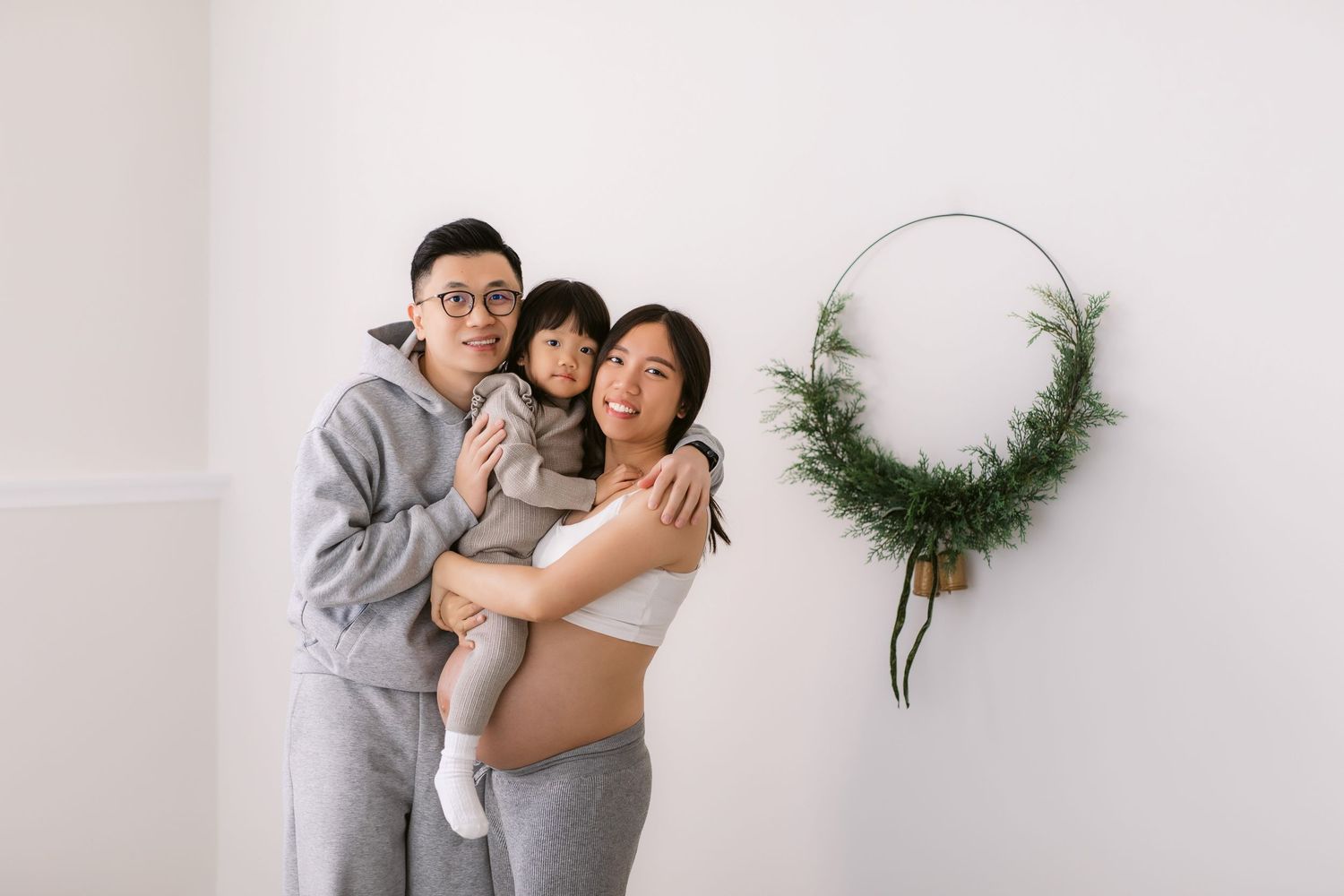 A family wearing matching gray loungewear poses together against a white wall with a minimalist greenery wreath decoration.