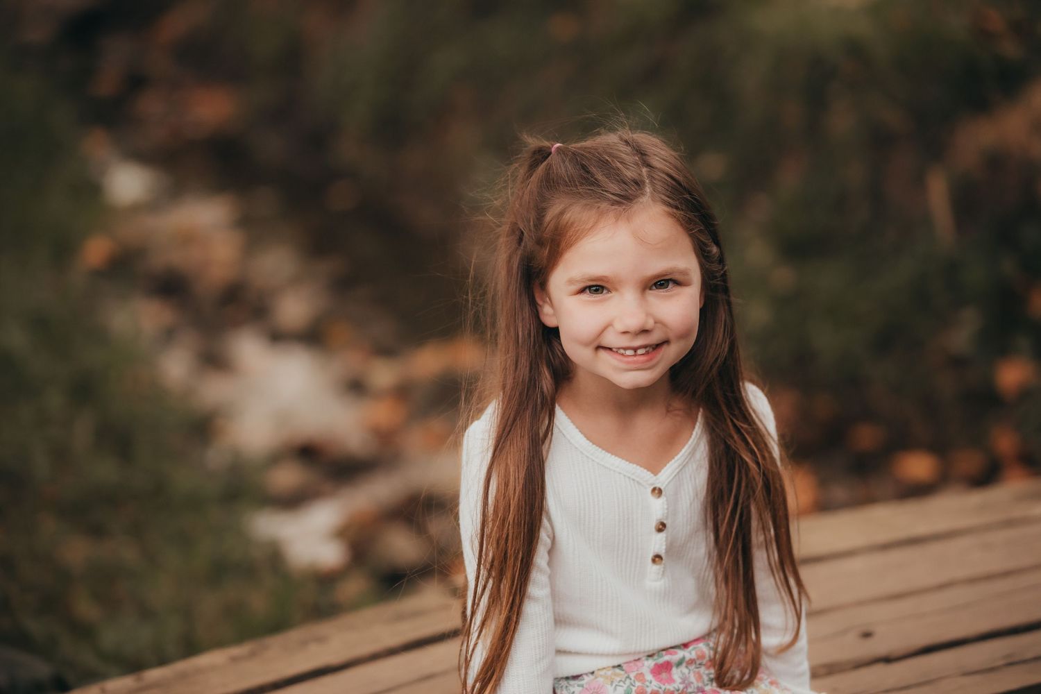 A child with long brown hair smiles warmly while sitting on wooden steps surrounded by autumn leaves.