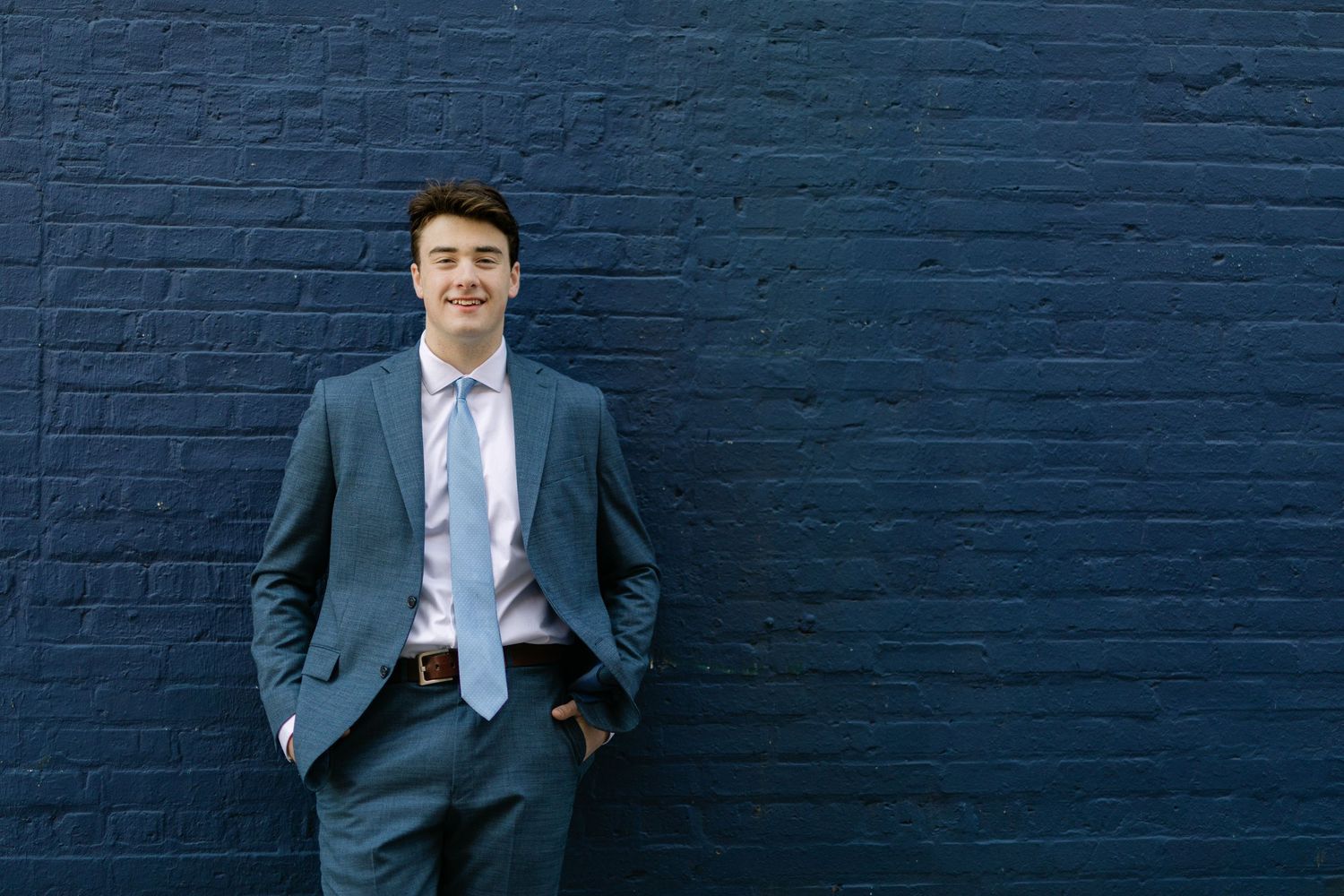 Young businessman in blue suit and tie smiling confidently against dark navy brick wall with hands in pockets.