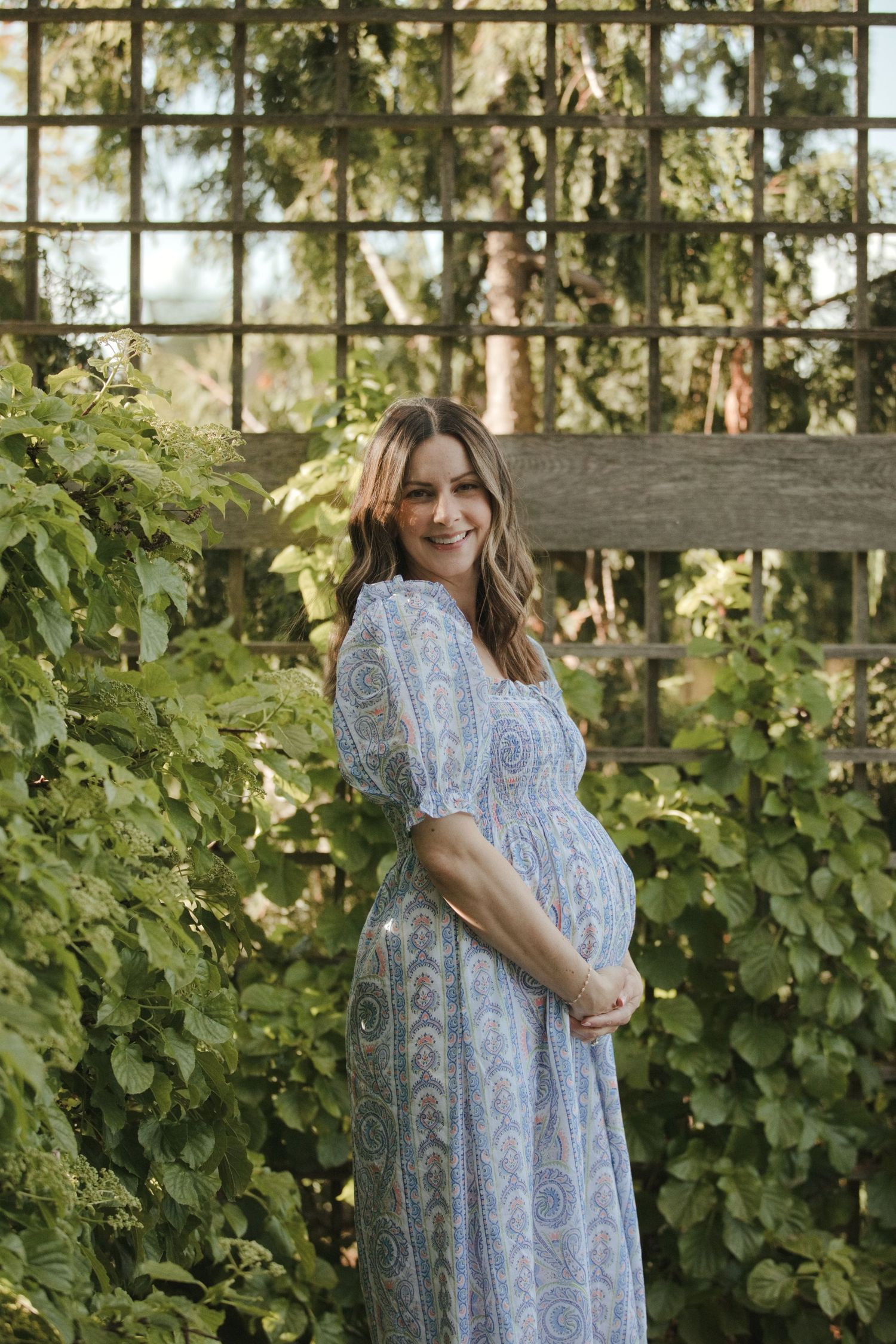 Smiling pregnant woman in blue floral maxi dress poses outdoors among lush green vines near wooden trellis.