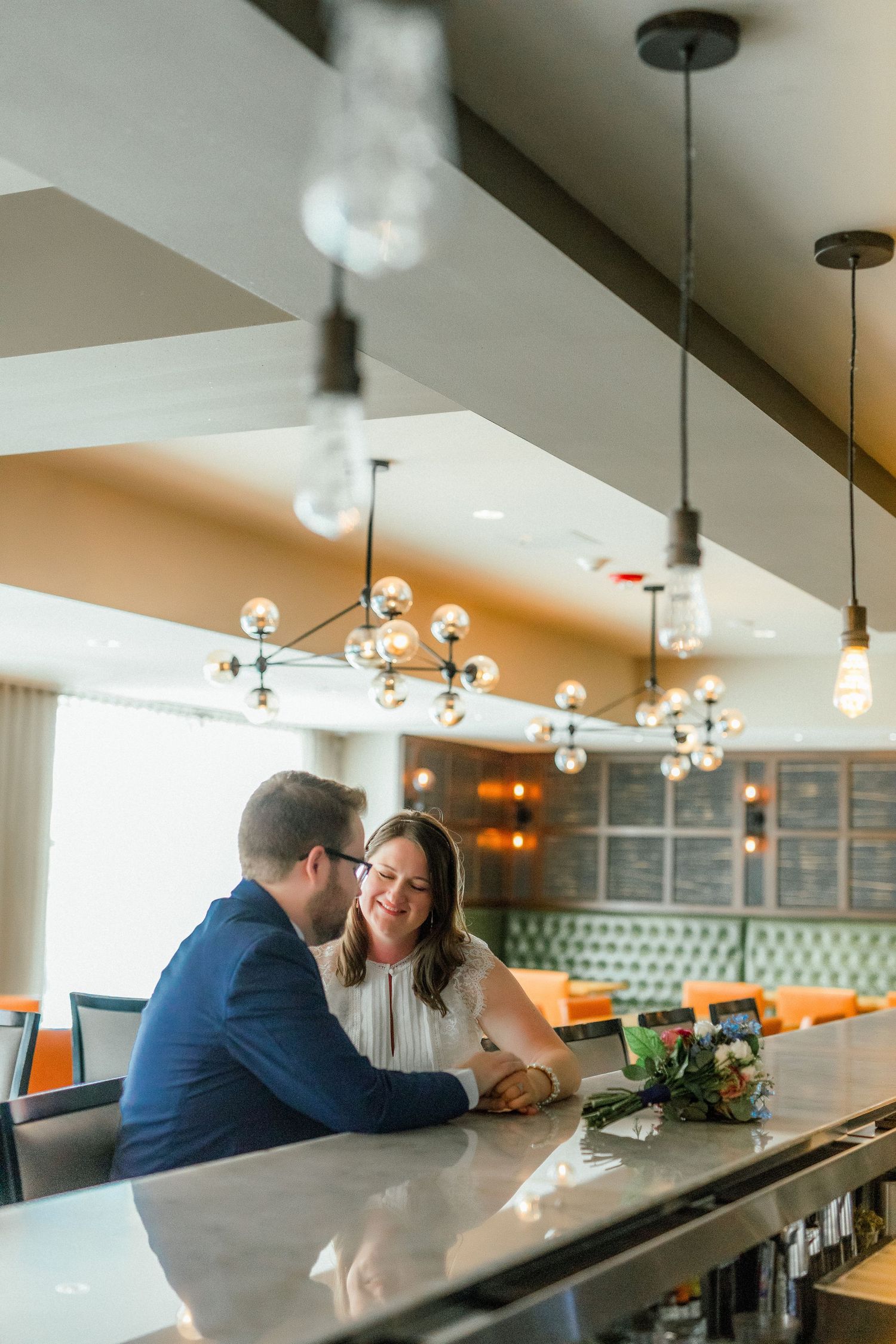 Couple smiling and holding hands at a modern restaurant bar with elegant pendant lights and colorful booth seating.