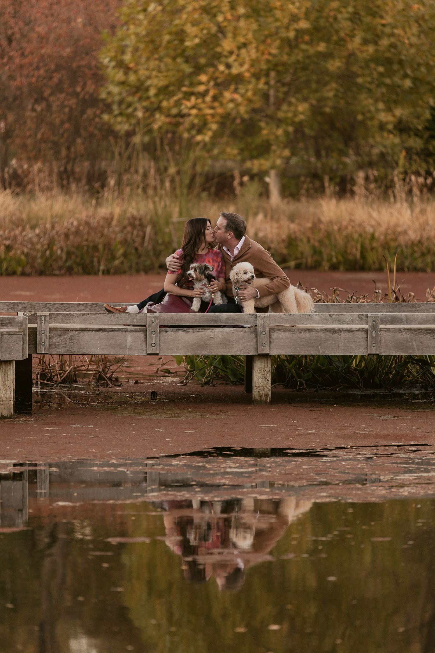 Romantic couple relaxing on wooden dock by pond during golden hour with autumn foliage in background.