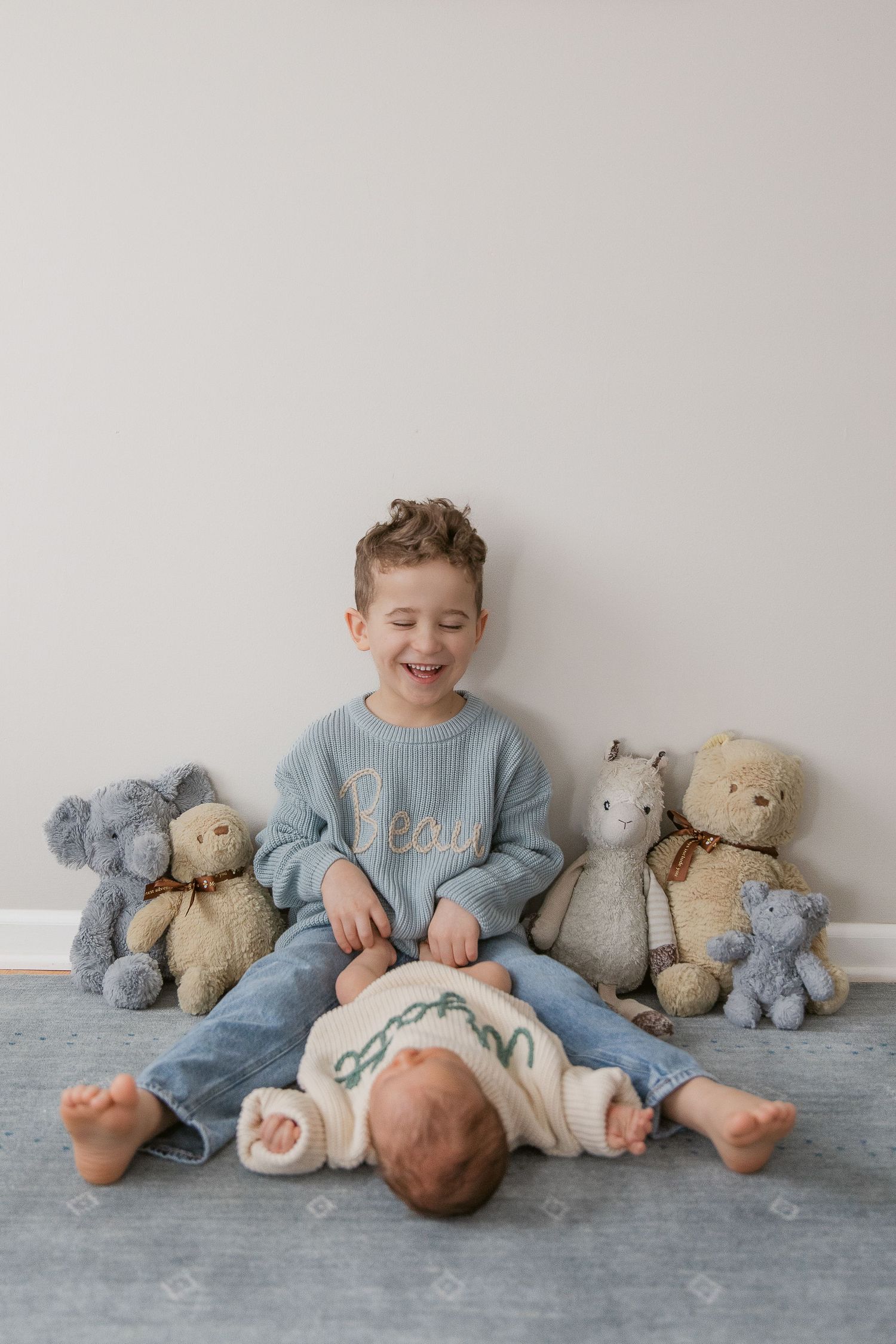 Siblings sitting against white wall with stuffed animal toys in blue and neutral tones.