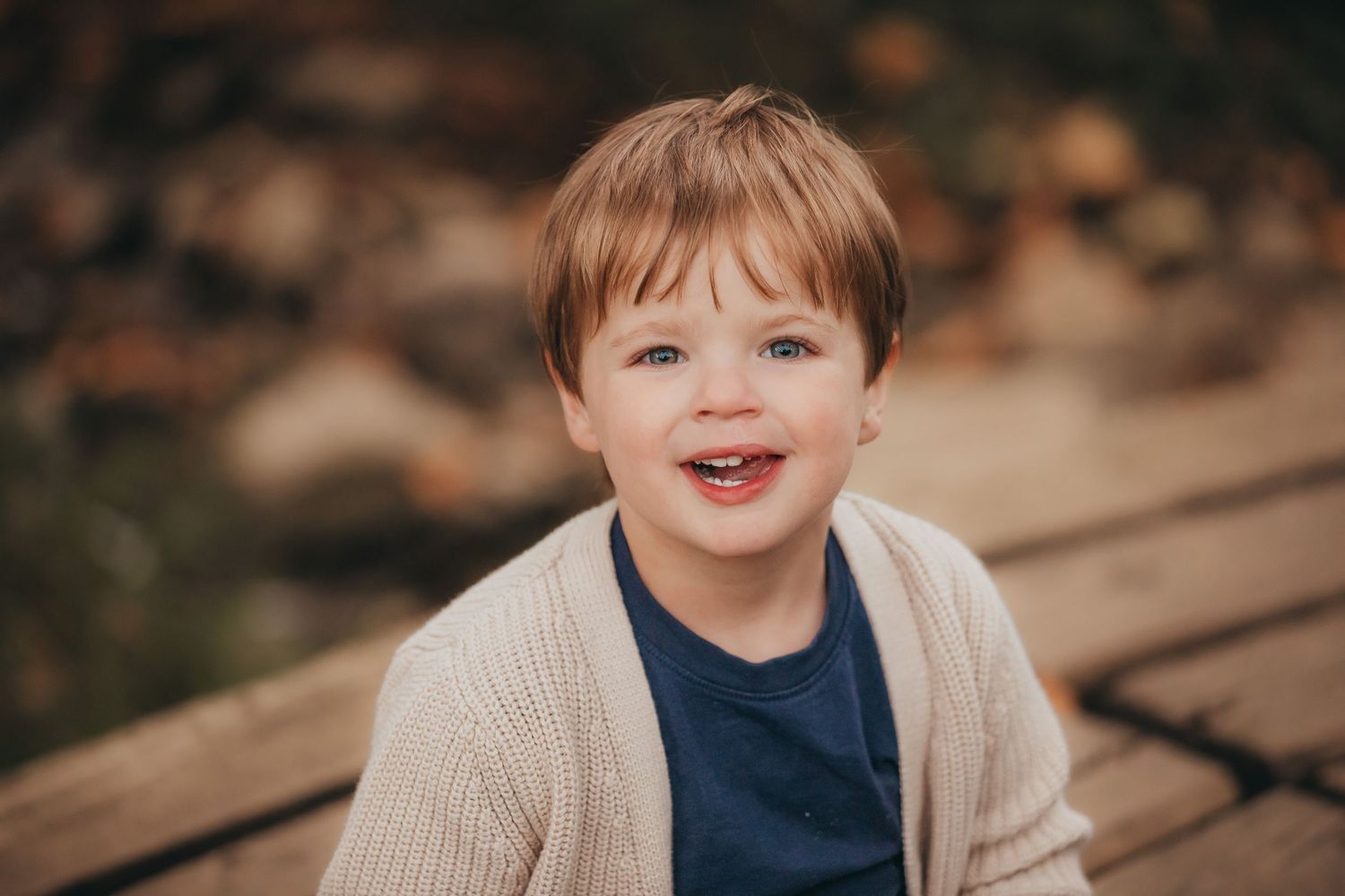 A seated toddler wearing a beige cardigan and navy shirt smiles joyfully in front of a blurred autumn background.
