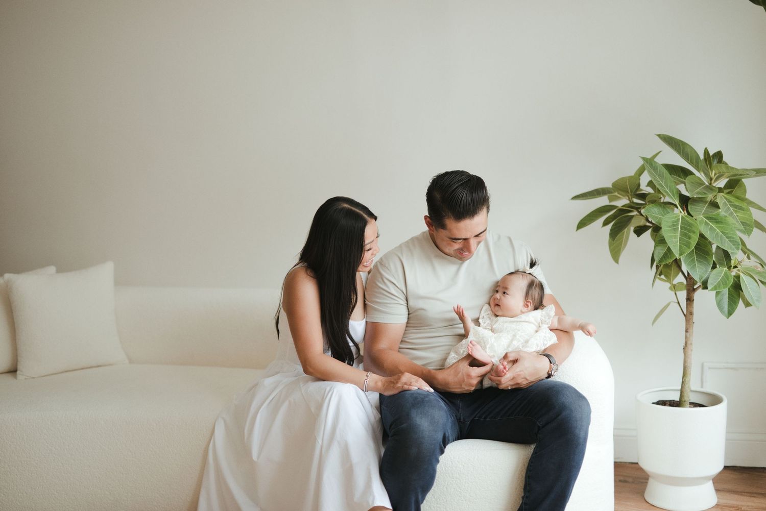 Young parents sitting on white sofa admiring their newborn baby in a bright, modern living room.