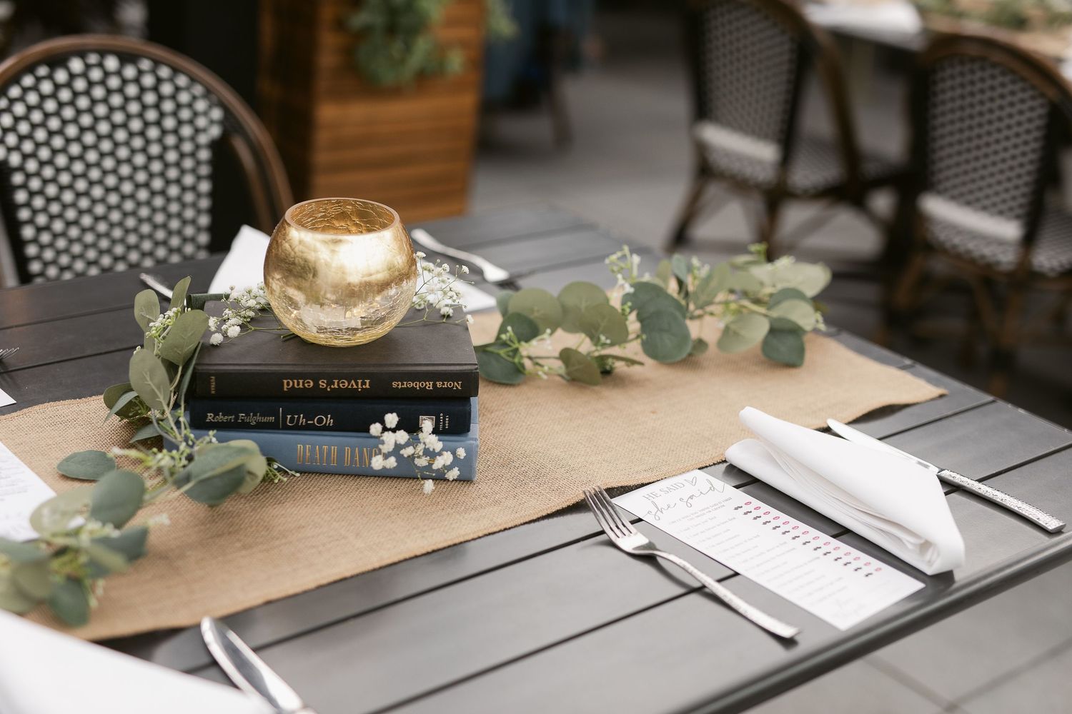 Elegant outdoor table setting with eucalyptus garland, stacked books, gold candle, and burlap runner for a wedding reception.