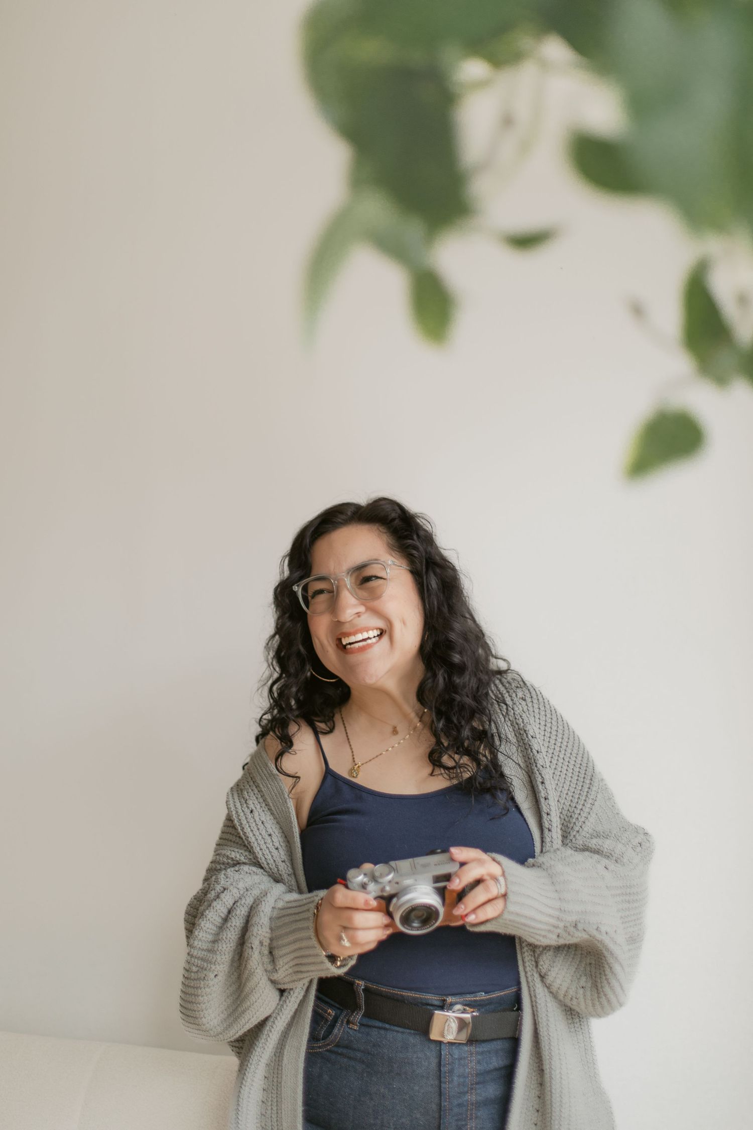 A person in a gray cardigan and blue top laughs joyfully while holding glasses against a light background with plants.