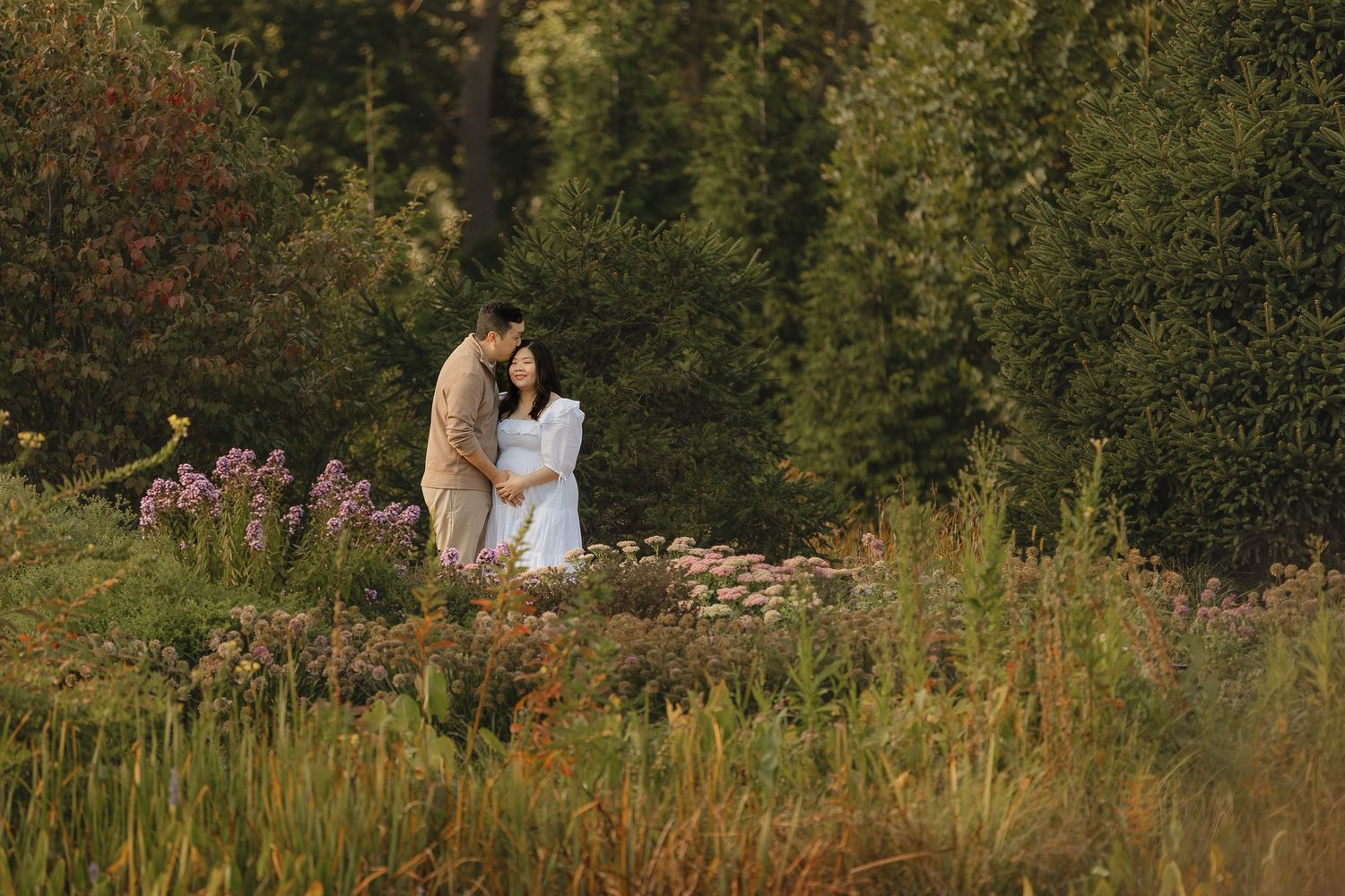 A couple stands together on a rustic stone wall surrounded by lush summer greenery and wildflowers at sunset.