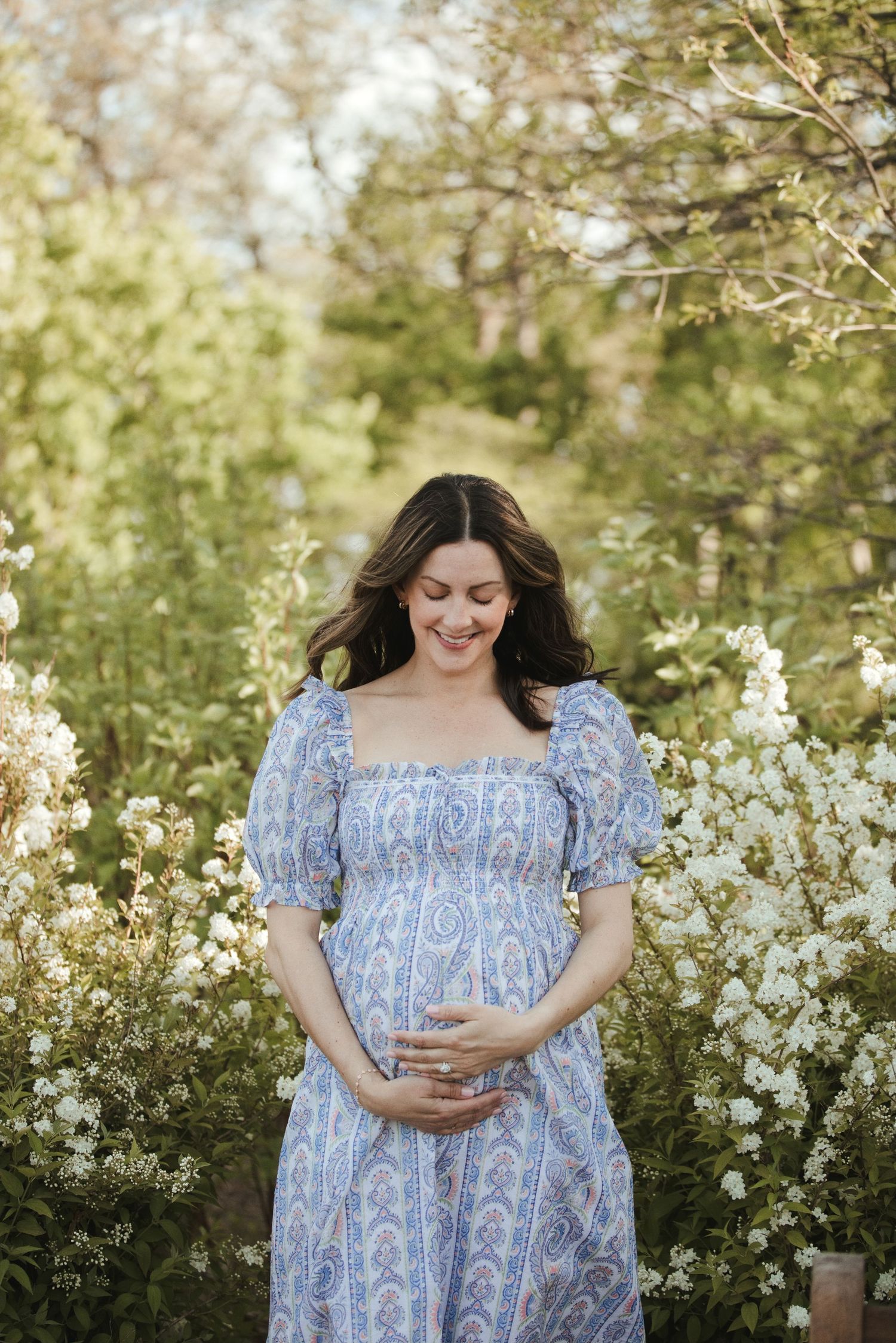 Smiling pregnant woman in blue floral dress cradling baby bump among white blooming flowers outdoors.