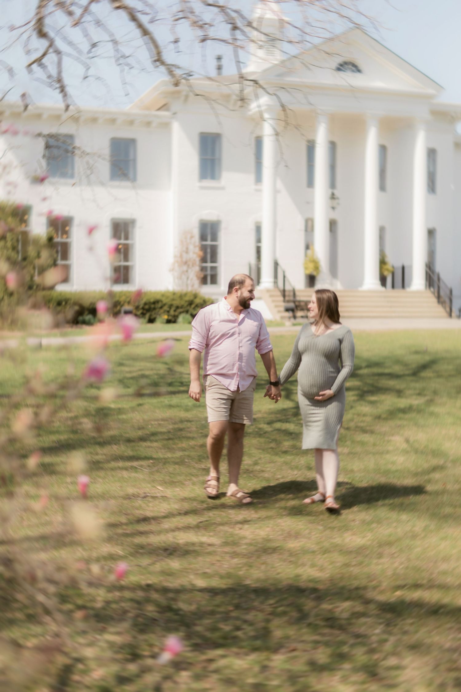A couple strolls on the lawn of a stately white columned mansion with spring blossoms in the foreground.