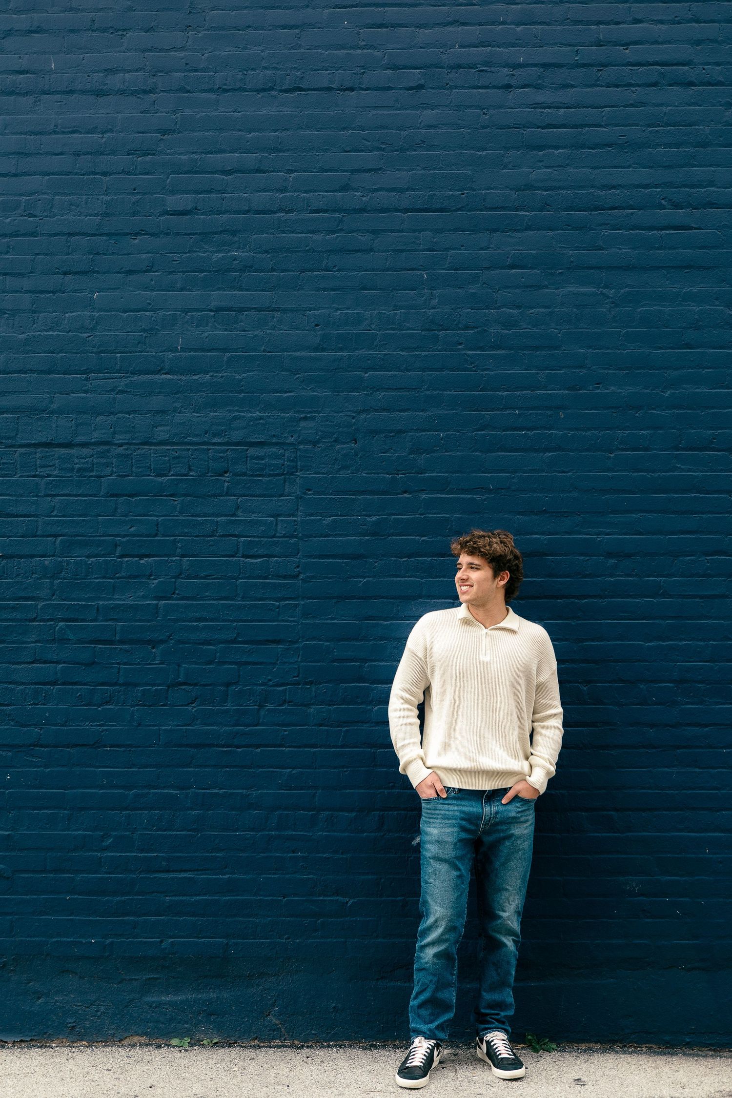 Person in cream sweater and jeans poses casually against navy blue brick wall.