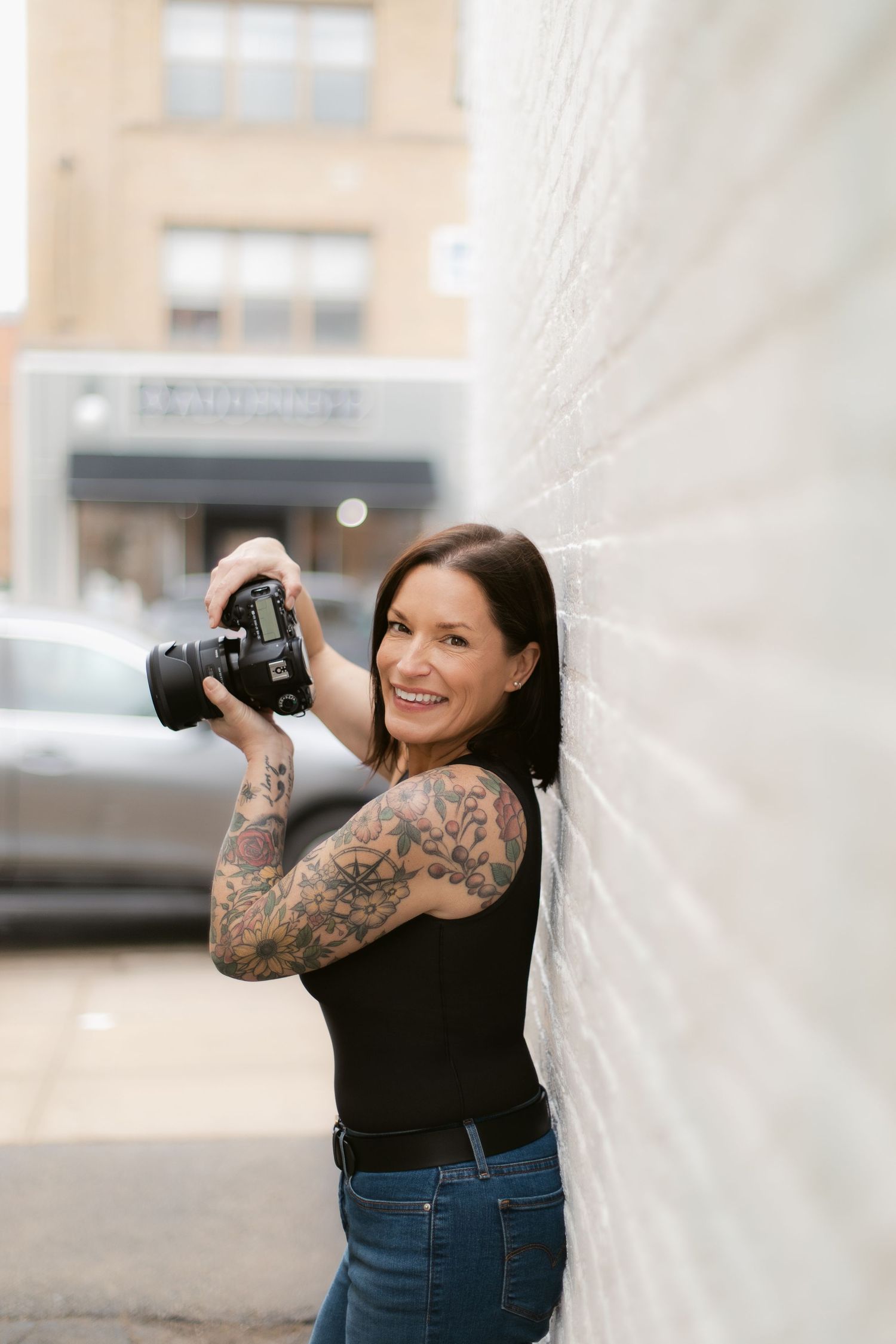 Smiling woman with tattoos leaning against white brick wall holding DSLR camera in urban setting.