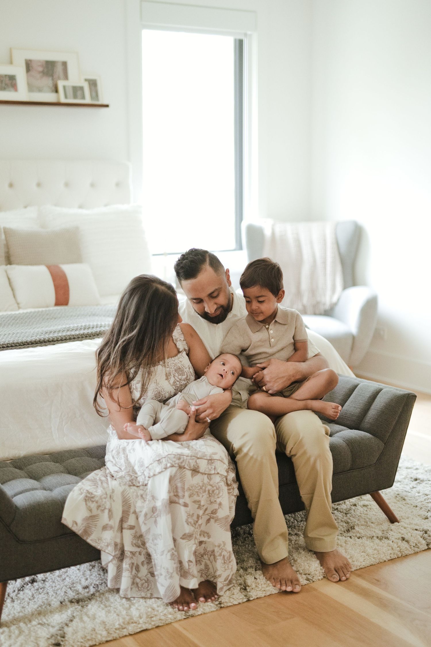A father sits on a bench holding a newborn baby while his daughter and son gather around him in a bright bedroom.
