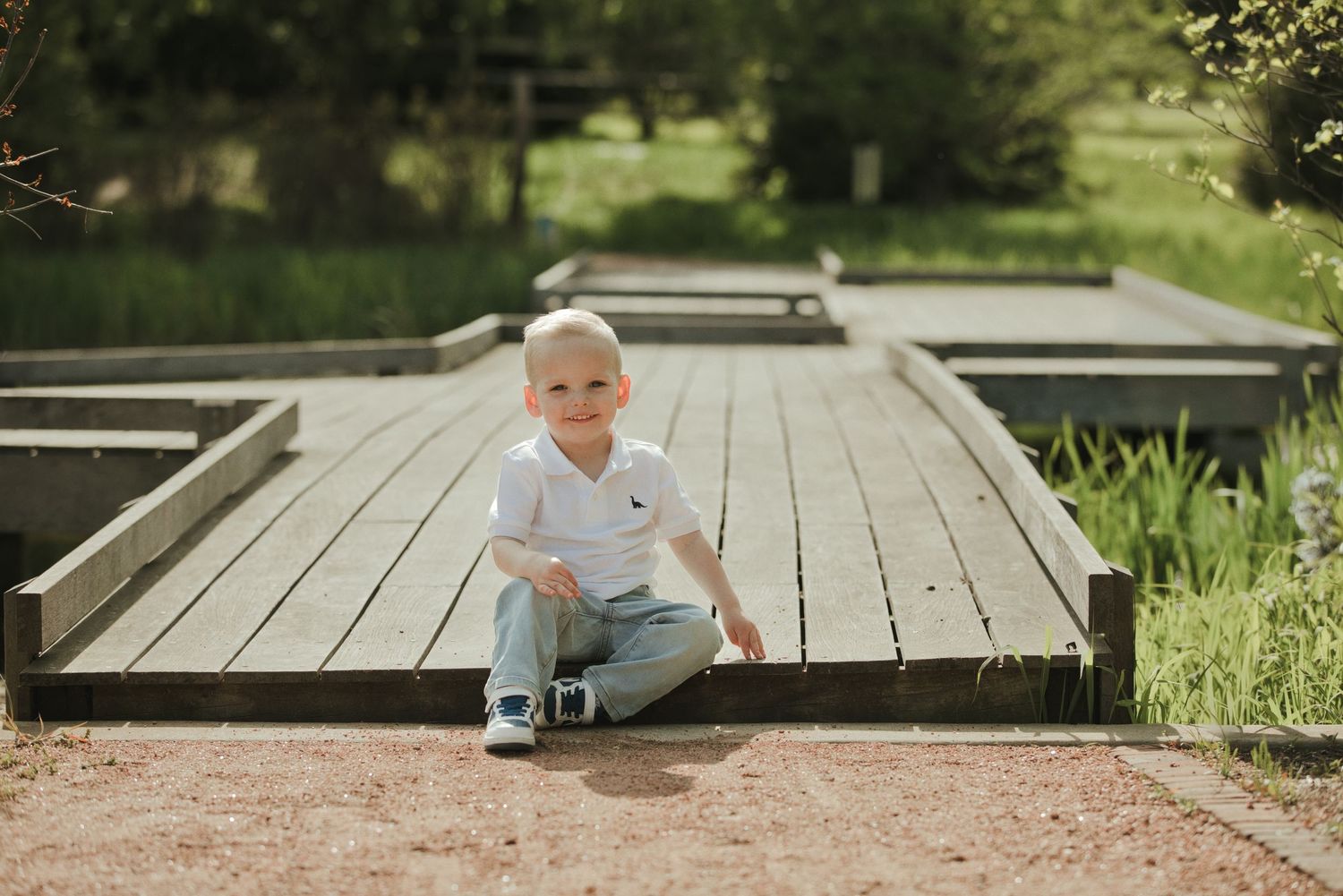 Smiling toddler boy in white shirt sitting on a wooden deck bridge surrounded by lush greenery outdoors.