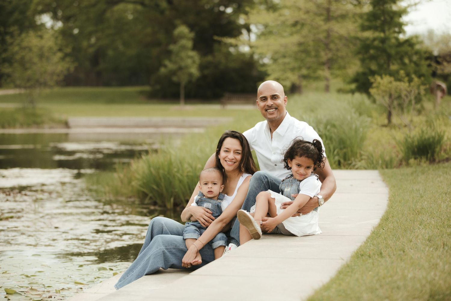 Happy family of four sitting together by a serene pond in a lush green park on a warm day.