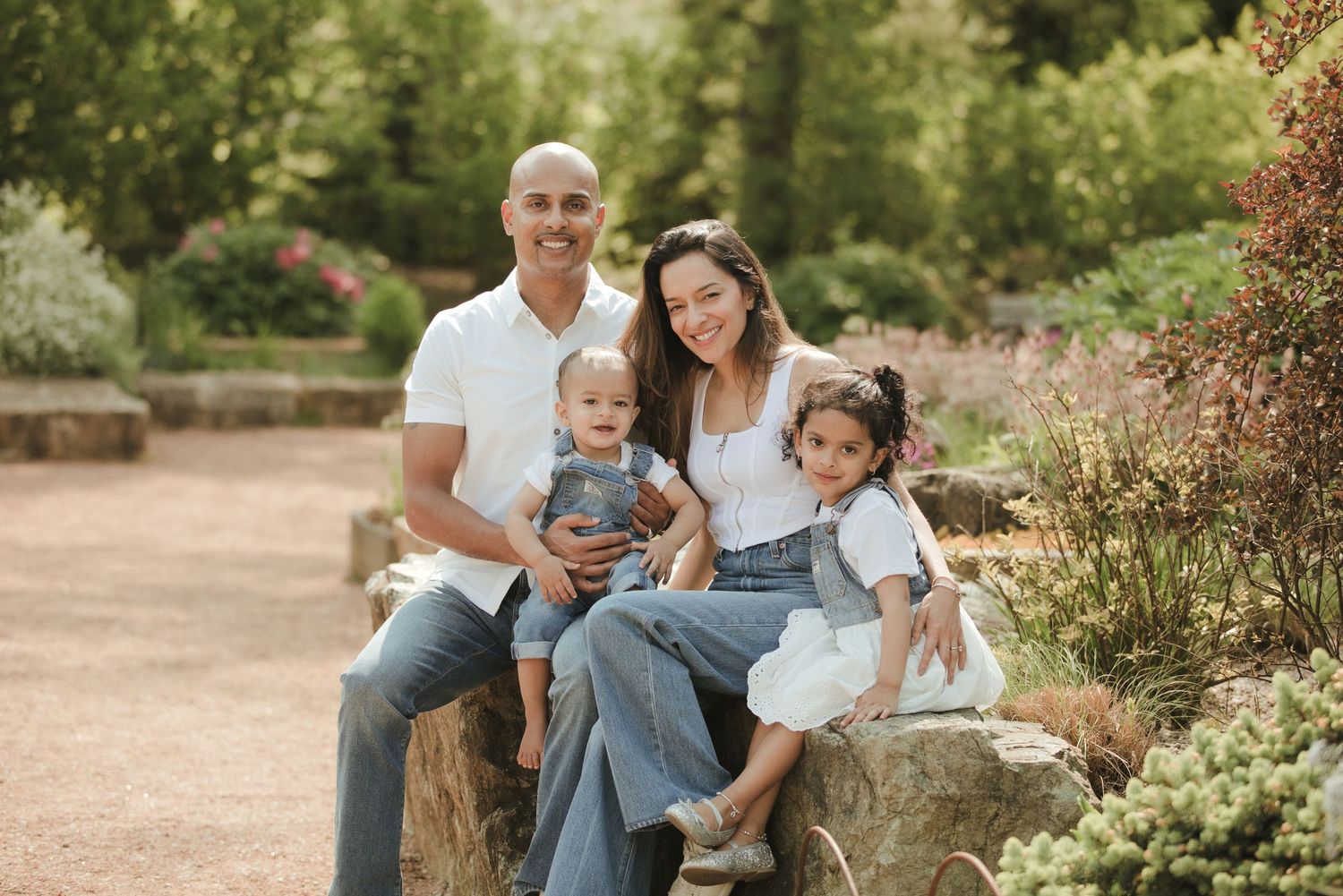 Family of four posing outdoors in a lush garden setting, parents and two young children smiling on a stone bench.